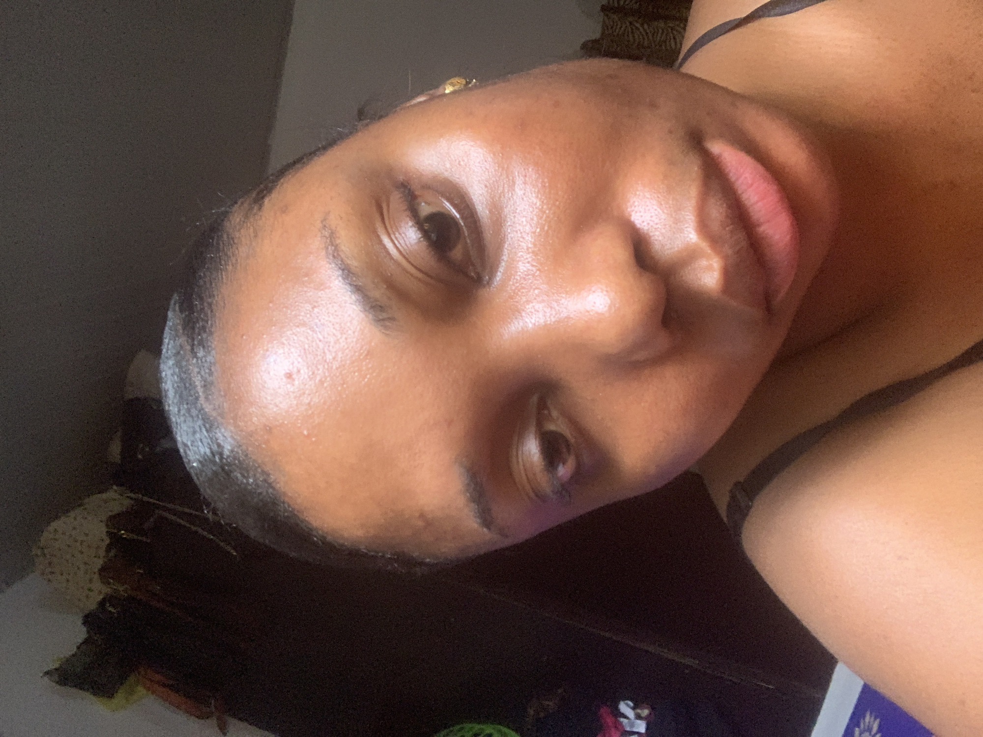 A close-up photo shows a dark-skinned woman with shiny skin. She has dark eyes and full, pink lips. She is wearing a thin-strapped top. The background shows a dark wooden surface and a bit of a gray wall. There is no verifiable information in the image to determine that the location is Abuja, Nigeria.