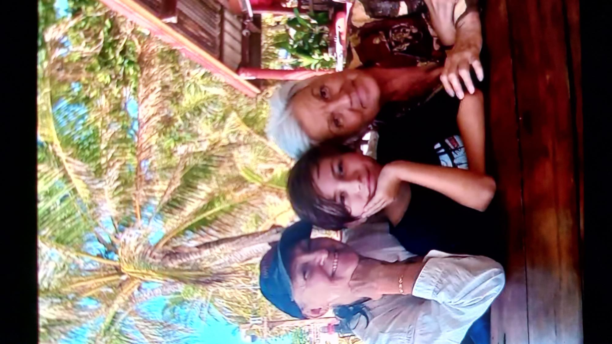 The image shows a medium shot of three people posing for a photo, likely at an outdoor cafe or restaurant. In the foreground, a young person with dark hair and a black t-shirt is positioned between two older women. The person on the left is wearing a blue baseball cap and a white shirt, with their hand resting on their chin. The person on the right has light gray hair and is wearing a patterned shirt, with their arm draped over the young person. They are all smiling and looking towards the camera. The background is blurred, but suggests a tropical setting with palm trees and what appears to be a roof structure. The location is identified as ตำบลเกาะพะงัน, Thailand.