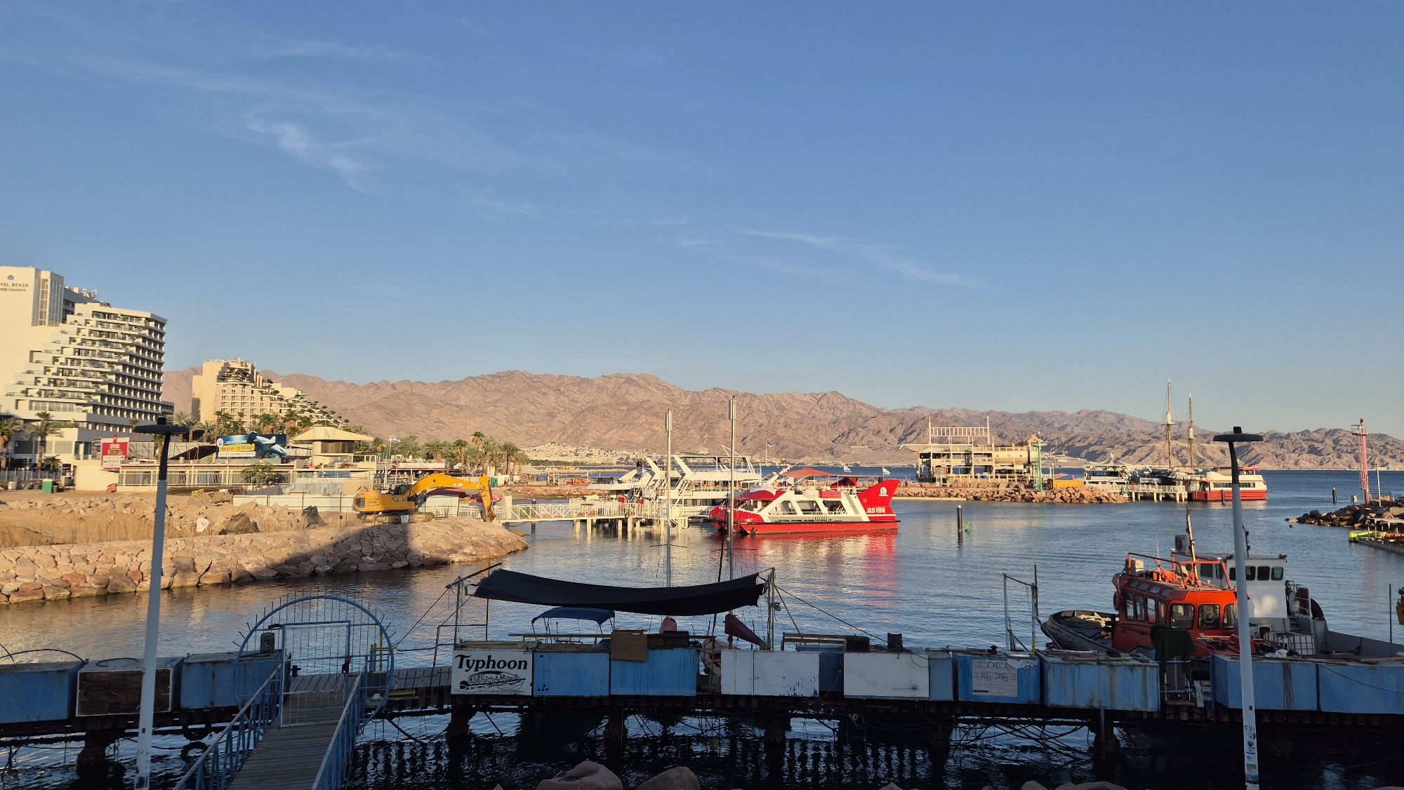 The photograph depicts a harbor scene in Eilat, Israel, during daylight hours. The foreground shows a weathered dock with a partially obscured blue-and-white structure bearing the word "Typhoon." A red and orange workboat is moored adjacent to the dock. The midground features a harbor with calm water reflecting sunlight. Several boats and yachts are docked along a stone breakwater. A red double-decker tour boat named "JULES VERNE" is prominently visible. In the background, there is a cityscape with tall buildings including the "Royal Beach Hotel," and hillside structures. Mountains rise behind the city. An excavator is visible on the breakwater. The sky is clear and blue with a few wispy clouds.