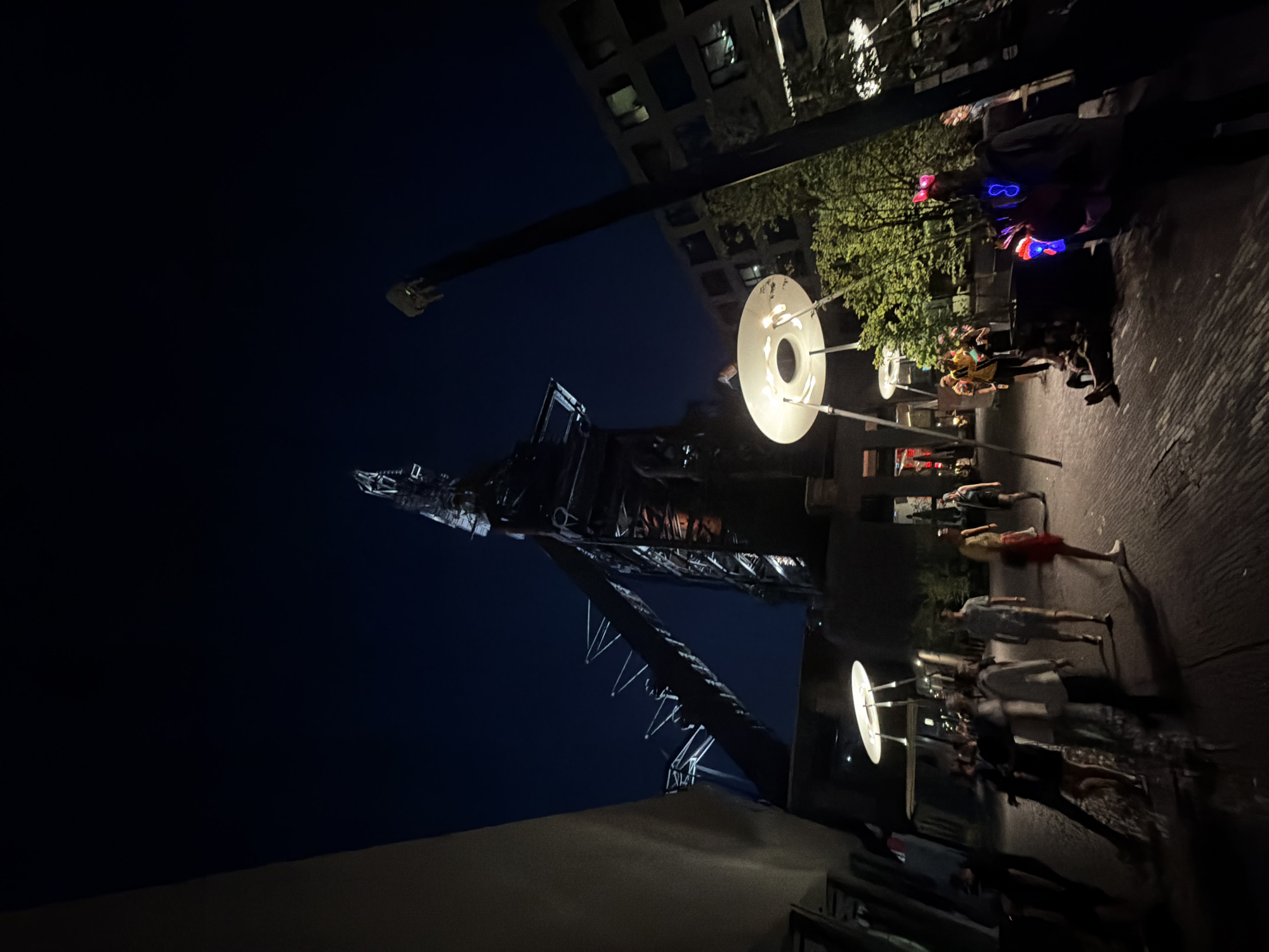 This is a low-angle, nighttime shot of the Belval blast furnaces in Esch-sur-Alzette, Luxembourg. The shot is taken from street level, looking up at the structure. In the right portion of the frame, there is a well-lit area populated with pedestrians. Some are wearing illuminated accessories, like glasses. There are decorative, disc-shaped lights on poles among the people. The building’s infrastructure, including the tower, is a prominent feature. The background sky is dark, indicative of night. Some modern buildings can be seen in the background.