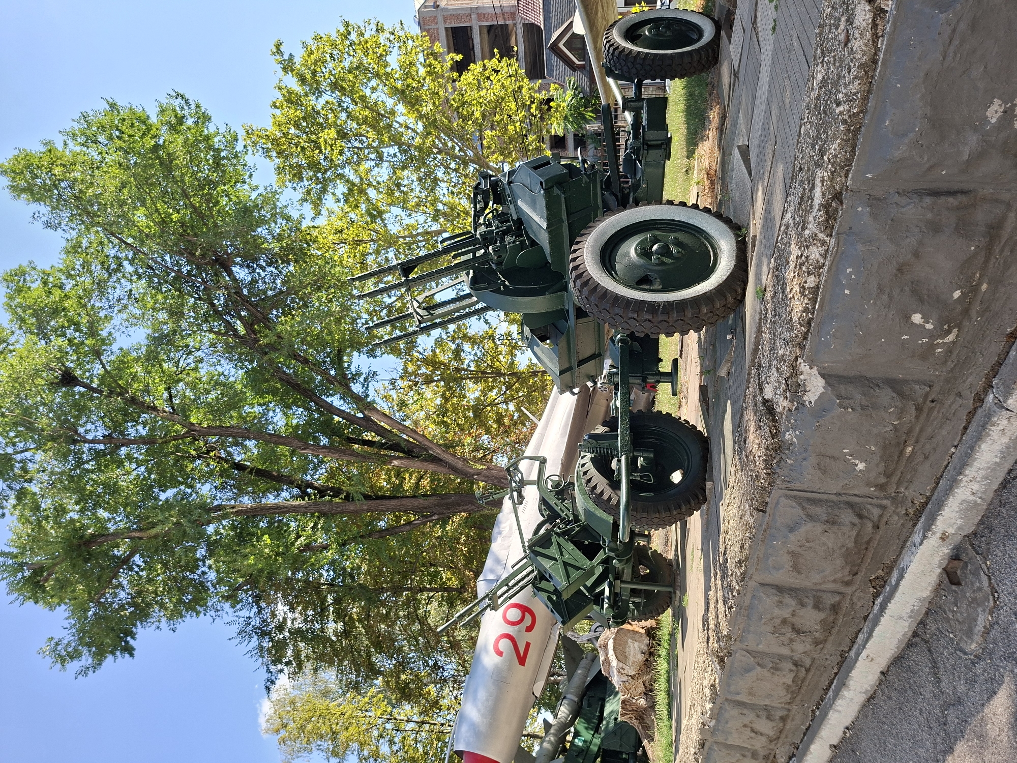 The image shows a static display of military hardware, likely in an outdoor museum or memorial site, with a clear blue sky and green trees in the background. The primary subjects are a large, silver missile with red markings, specifically the number "29", and a green, wheeled anti-aircraft gun with multiple barrels. The missile is positioned horizontally, resting on a support structure. The anti-aircraft gun is situated in front of and partially beneath the missile, with its wheels on a paved surface. Behind these objects, a building with visible construction elements and some residential buildings are present, suggesting an urban or semi-urban environment. The foreground features a textured concrete wall and curb, with paved ground. The scene is brightly lit by sunlight. Based on publicly available information, this display is located at the Military Glory Memorial (Memorialul Gloriei Militare) in Chișinău, Moldova. No people are visible in the image.