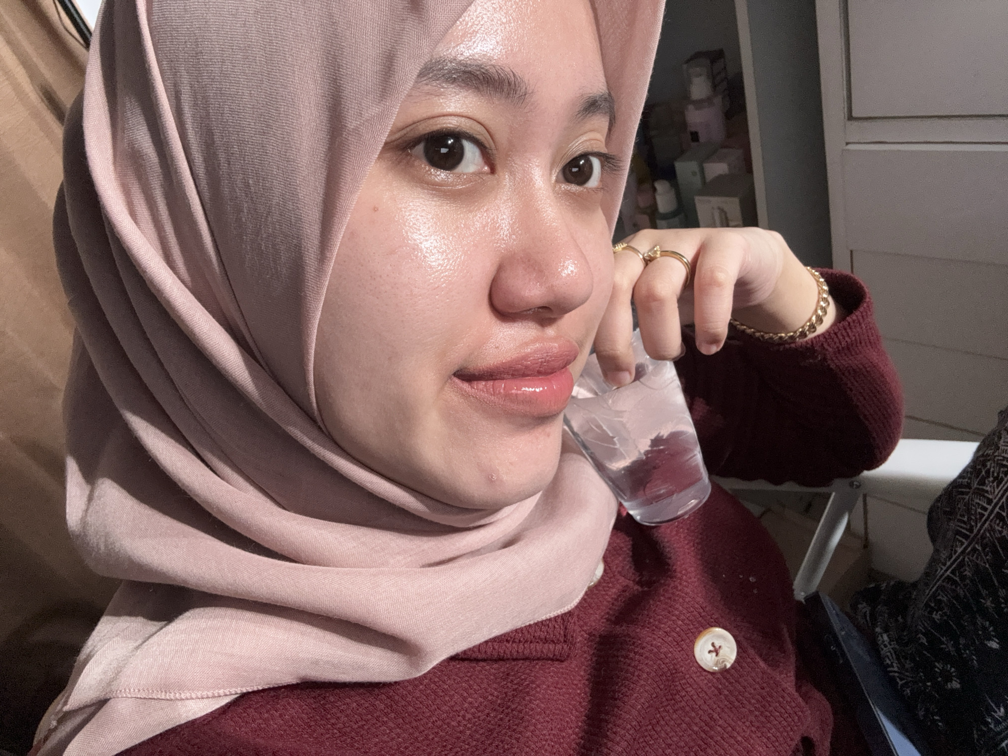 A person wearing a pink hijab and a maroon knit sweater is holding a clear glass of water. The person is wearing gold jewelry, including rings and a bracelet. A button is visible on the sweater. Behind the person is a white dresser with various items on top. The location is Gadingrejo, Indonesia.
