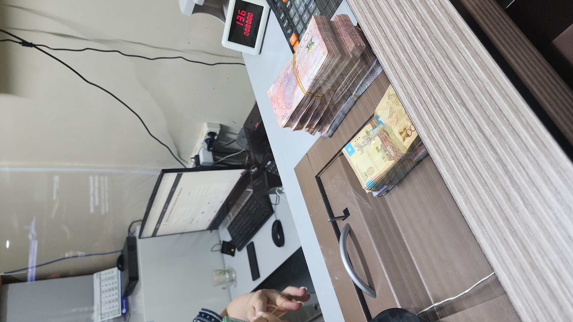 The image depicts a transaction counter, likely within a financial institution or exchange office in Shymkent, Kazakhstan. A stack of banknotes, appearing to be tenge, is prominently displayed on the counter. These notes are bundled with elastic bands, suggesting they are either being counted or prepared for a transaction.
On the left side of the counter, a person's hands are visible, engaged in handling more banknotes. This action further reinforces the context of a financial transaction. Behind the counter, a computer monitor displays a screen with what seems to be a financial application or system. A keyboard and mouse are positioned in front of the monitor, indicating the presence of a workstation.
To the right of the banknotes, a digital counter shows "136" on the top line and "000000" on the bottom line, possibly indicating a quantity or a code related to the transaction. A calculator is also visible on the counter. A calendar is placed on a shelf in the background. The environment appears to be an indoor setting with artificial lighting, and the overall impression is one of a service area where financial exchanges take place.