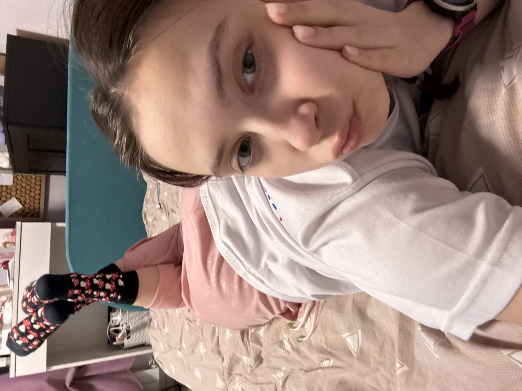 A young person is lying on a bed with a light beige patterned comforter. They are wearing a white t-shirt, pink pants, and black socks with red and white mushroom patterns. The person is holding their hand to their face. A dark smart watch is visible on the wrist. The room includes a teal chair, white shelving unit with items on it, and a dark dresser. Location cannot be determined.