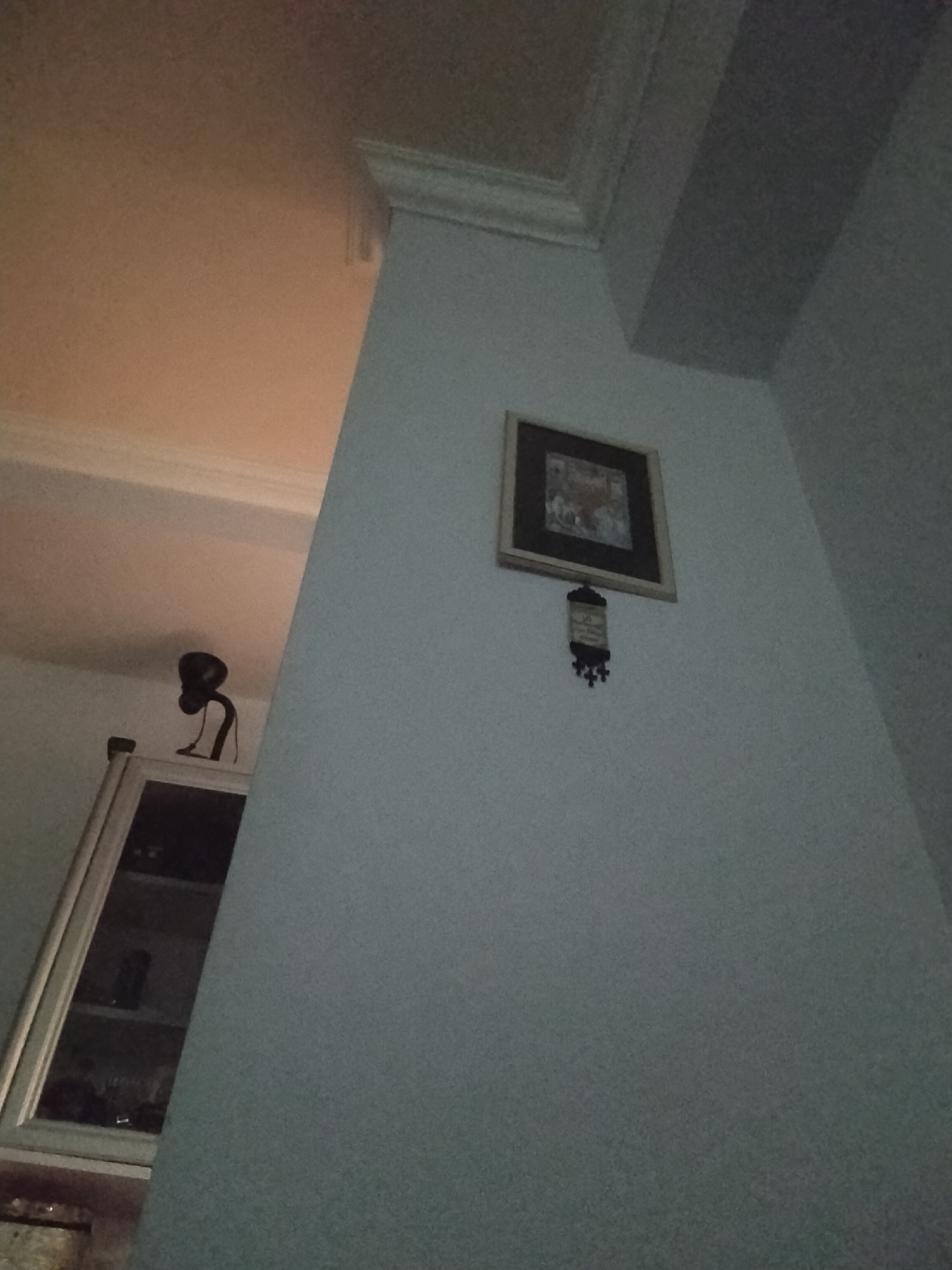 The image shows an interior corner of a room. The walls are light gray. A framed picture hangs on one wall, with a decorative item below it. A white cabinet with glass doors is visible in the background, with objects inside. A black lamp sits atop the cabinet. The location is inside a building, possibly a home, but the exact location in Yerevan, Armenia, cannot be determined from this image alone. No people or specific actions are visible.