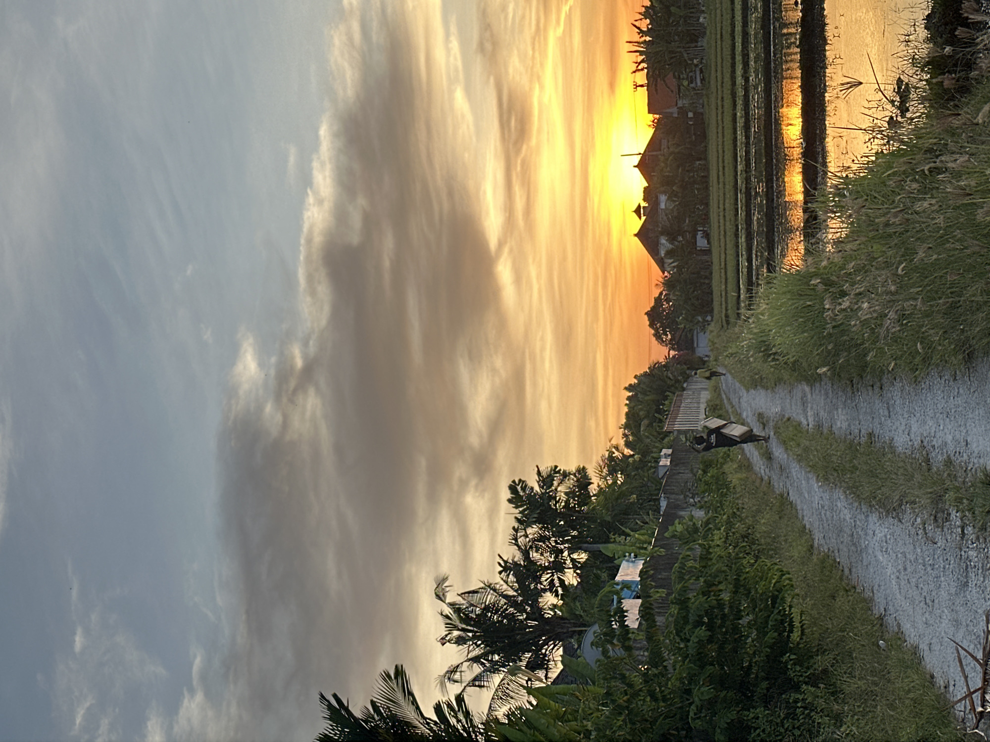 The image depicts a sunset scene in North Kuta, Indonesia. The sky is a mix of pale blue and orange hues, with clouds obscuring the setting sun. Below the horizon are rice paddies reflecting the sunlight, next to traditional Indonesian houses. A dirt path runs along the rice paddies, flanked by vegetation.