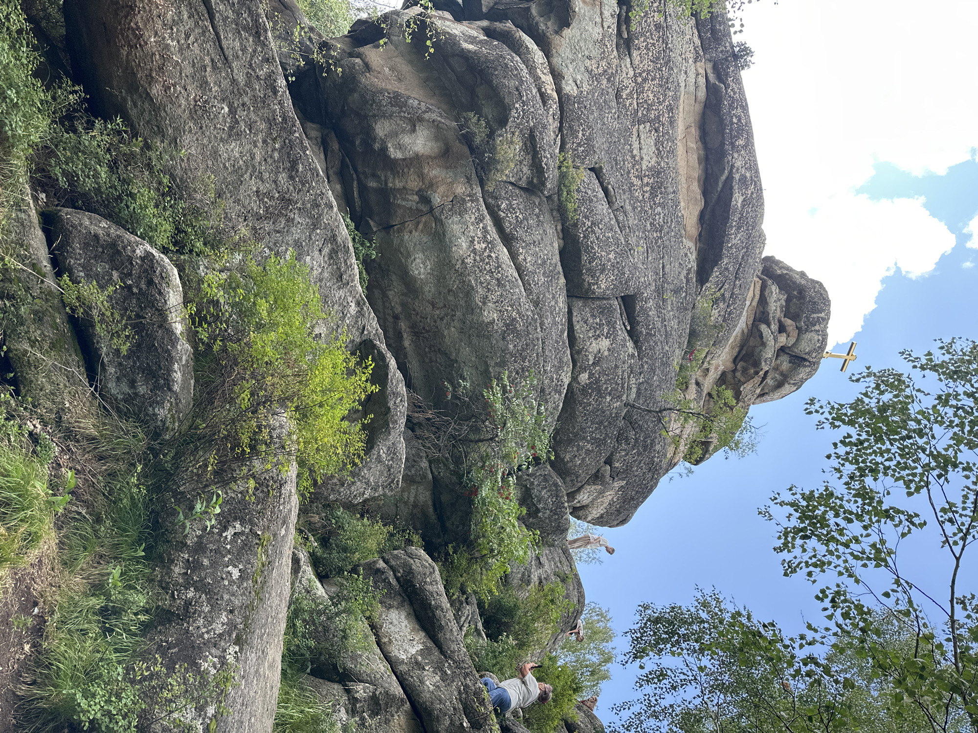 The image depicts a rocky, natural landscape with several large boulders. A golden cross is visible atop one of the highest rock formations. Three individuals are present: one person is standing on a rock, another is partially visible sitting on a rock, and a third person is in the foreground, holding a phone and seemingly taking a picture. The environment is characterized by lush green foliage, including trees and bushes, growing among and around the rocks. The sky is blue with scattered white clouds. The scene appears to be an outdoor tourist or hiking location.