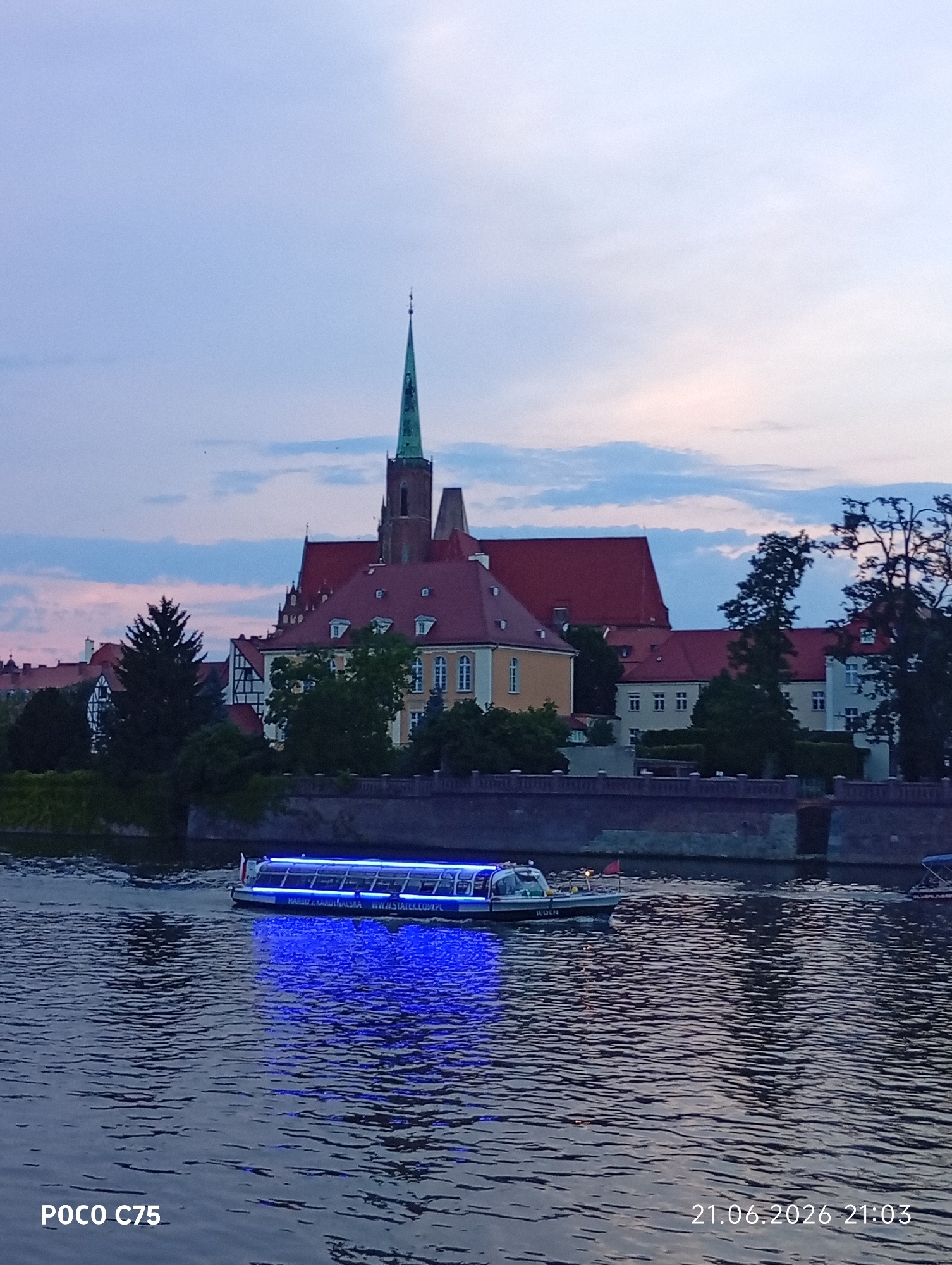 A boat with blue lights is cruising on a river in front of a cityscape. The boat appears to be a passenger vessel with a roof and windows, and it has text on its side that reads "HARBOUR KARDYNALSKA WWW.STATER.COM.PL". In the background, there is a large building complex with red roofs, characteristic of a historic European city. A prominent green spire, part of a church or cathedral, rises above the buildings. The sky is a soft gradient of blue and pink, suggesting it is either early morning or late evening. The river reflects the lights from the boat and the sky, creating a shimmering effect on the water. The overall scene depicts a tranquil view of a European city at dusk, with the river and a boat as the primary focus. The image is located in Wrocław, Poland, specifically along the Odra River with a view of Ostrów Tumski, the historic cathedral island, and the surrounding architecture.
