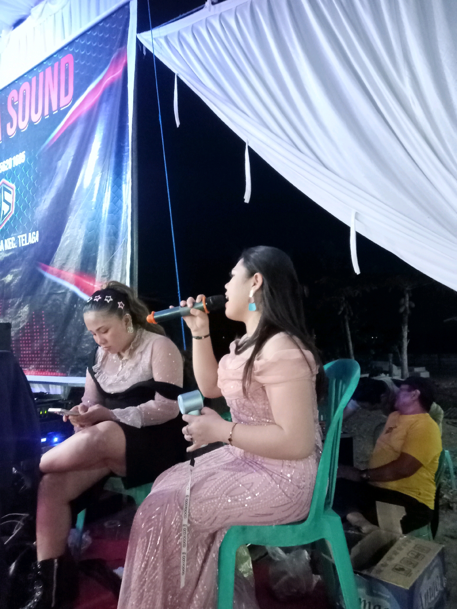 The image depicts an outdoor event at night, likely in Mongolato, Indonesia, suggested by the banner in the background which reads "SOUND" and mentions "KEC. TELAGA". The foreground features two women. The woman on the right is wearing a pink sequined dress and a light blue earring, holding a microphone, and appears to be singing. She is seated on a green plastic chair. The woman on the left is wearing a sheer black and pink dress with a sparkly overlay and has a star-shaped accessory in her hair. She is looking down at her phone, which she is holding in her hands. She is also seated. Behind them, partially visible, is a man in a yellow shirt. The environment appears to be a gathering or performance space, indicated by the stage setup with a banner and a tent-like white fabric overhead. Several objects are visible, including a microphone, a phone, chairs, and boxes on the ground. The lighting is dim, suggesting artificial illumination is in use.