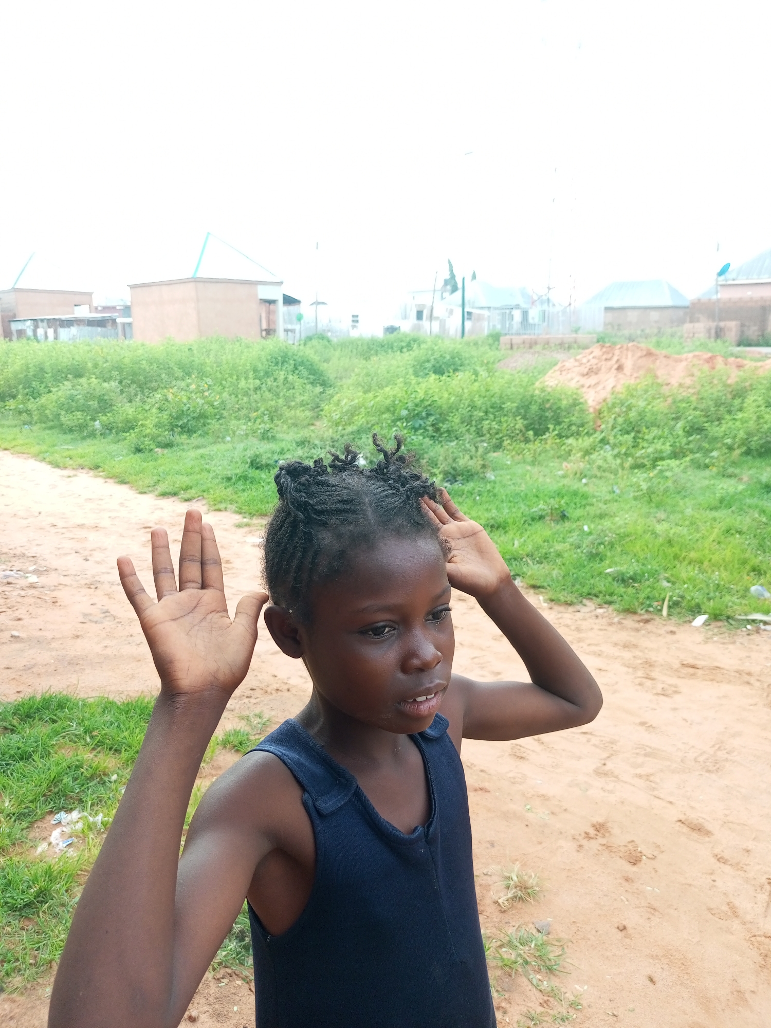 A young Black girl with braided hair, wearing a dark blue tank top, stands in a dusty outdoor area with green foliage and several low-rise buildings in the background. The girl's left hand is raised in front of her face, with her palm facing forward. Her right hand is touching her head. The environment appears to be a rural or semi-urban area within Tempure, Nigeria, characterized by dirt paths, sparse vegetation, and simple structures. The lighting is bright, suggesting it is daytime.