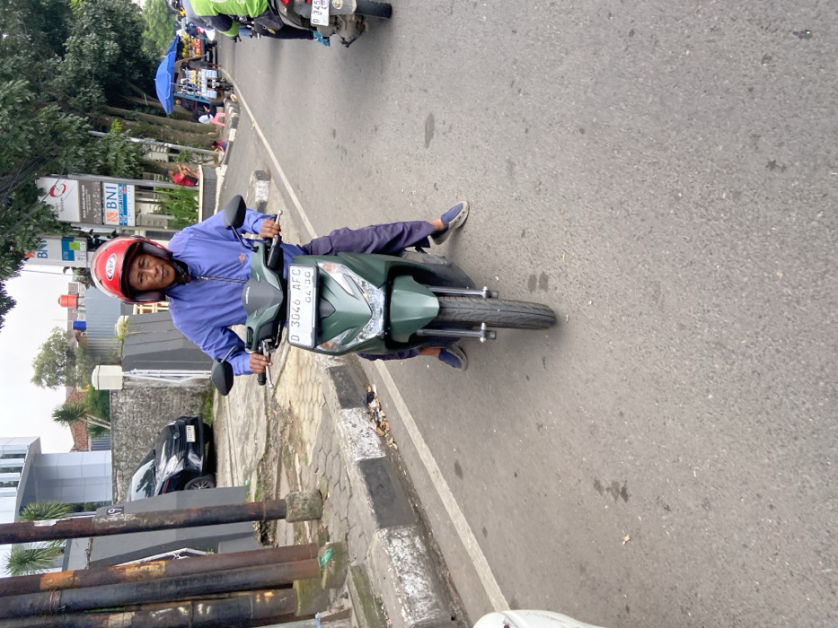 A man in a red helmet sits on a green scooter. The scooter's license plate reads "D 3046 AFC." The man is wearing a blue long-sleeved shirt, blue jeans, and black sandals. The man is facing forward. The scooter is stopped in the middle of a paved road with white lines on either side. The scooter is facing forward. There are several buildings in the background. The image is taken in Bandung, Indonesia, at coordinates (latitude: -6.9, longitude: 107.628).