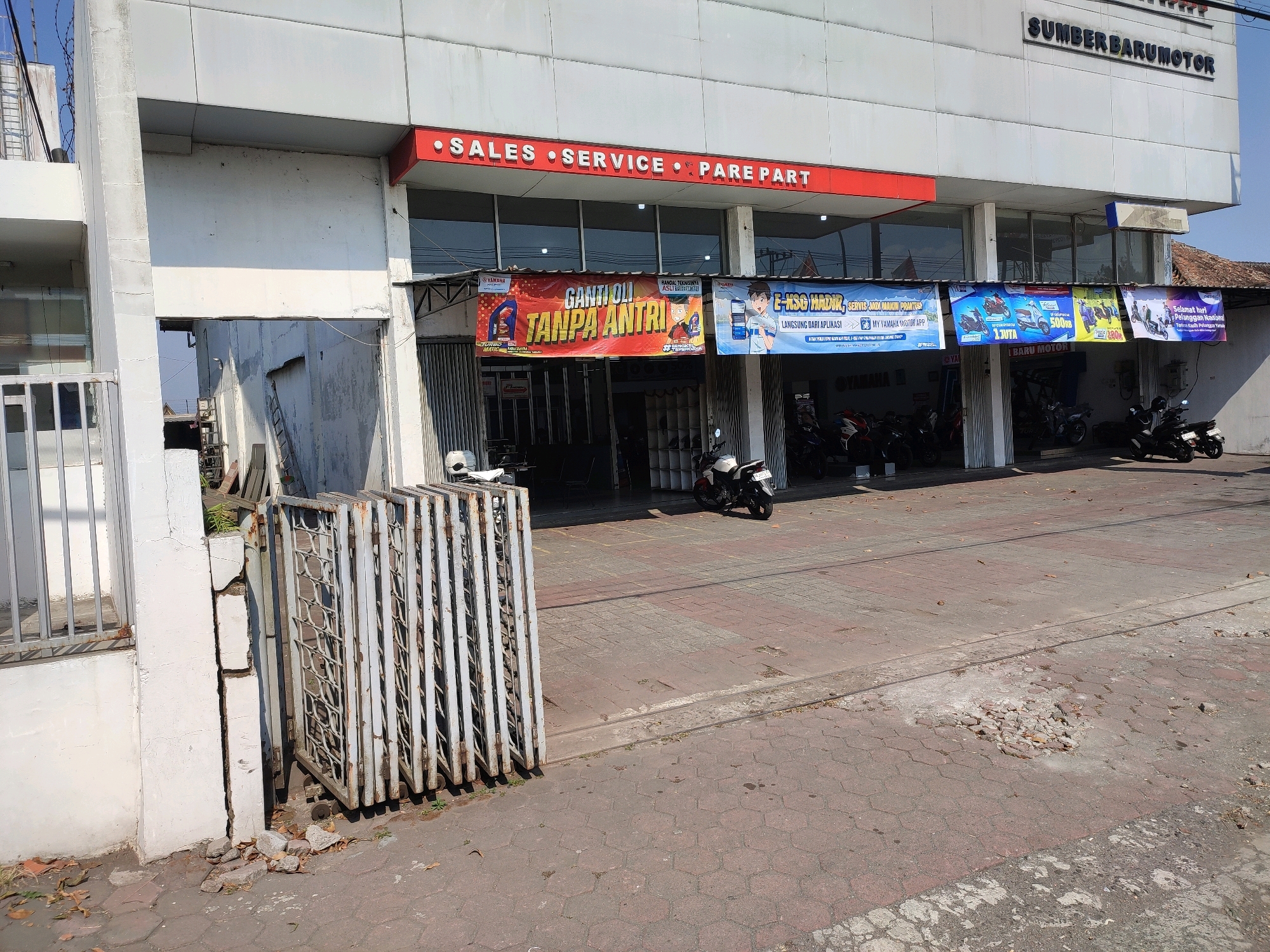The image shows the exterior of a motorcycle dealership called "SUMBER BARUMOTOR" located in Magelang, Indonesia. The building is white with a red awning above the entrance that reads "SALES • SERVICE • PARE PART". Several banners are displayed outside, promoting various services and offers, including "GANTI OLI TANPA ANTRI" (Oil Change Without Queuing) and an app called "MY YAMAHA MOTOR APP". Various Yamaha motorcycles are parked in front of the dealership, some under a covered area. The ground in front of the building is paved with interlocking bricks, and there are some damaged sections. A metal gate is visible on the left side of the image. The overall scene depicts a typical motorcycle dealership on a sunny day.