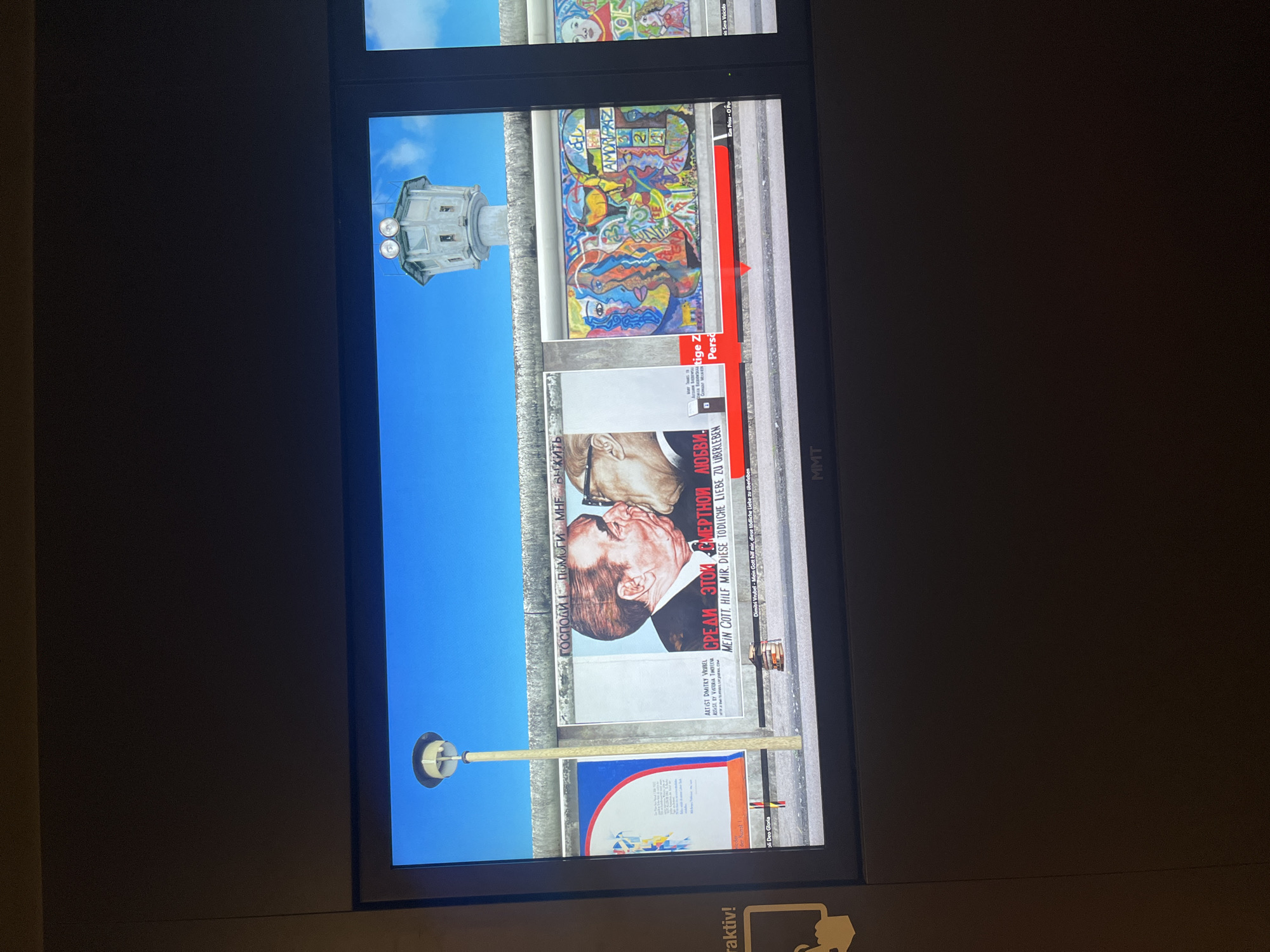 The image displays a digital reproduction of the East Side Gallery in Berlin, Germany. This is a section of the Berlin Wall that has been transformed into an open-air art gallery. The scene shows a vibrant blue sky above a graffiti-covered section of the wall, which features several distinct murals.
Prominently displayed is a well-known mural depicting a fraternal kiss between Leonid Brezhnev and Erich Honecker, leaders of the Soviet Union and East Germany respectively. Below this image, text in Russian and German reads "Gott, hilf mir, diese tödliche Liebe zu überleben," which translates to "God, help me survive this deadly love."
To the right of this mural is a colorful abstract artwork featuring stylized human faces and the word "AMOR-PAZ," meaning "Love-Peace." Further to the right, another mural shows a depiction of a young girl.
In the background, a concrete watchtower, a remnant of the wall's original purpose as a border fortification, stands against the sky. A tall, slender lamppost stands to the left of the murals. The bottom of the display shows the brand name "MMT." The overall impression is that of a historical exhibit, likely in a museum or gallery setting, showcasing a segment of the Berlin Wall's artistic transformation.