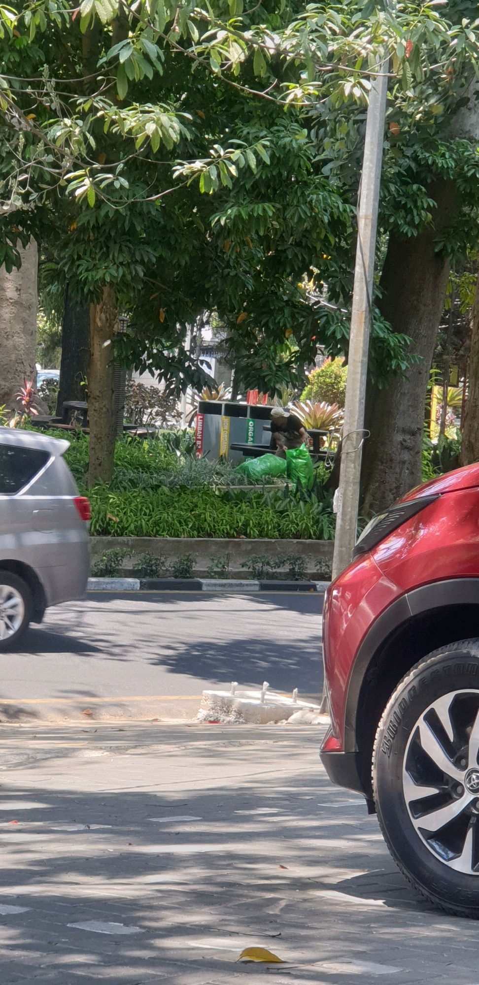 The image depicts a street scene in Bandung City, Indonesia. In the foreground, the front portion of a red car is visible on the right, and a silver car is on the left. Behind the cars, a person wearing a light-colored cap and a dark shirt is crouched near some green trash bags. The person appears to be interacting with the trash bags, possibly collecting or organizing them. Behind the person, there are several large trash receptacles with labels, and further back, lush green foliage and trees create a natural backdrop. The scene is illuminated by natural sunlight, casting shadows on the road and pavement. The environment suggests a public area, potentially a park or a sidewalk adjacent to a green space.