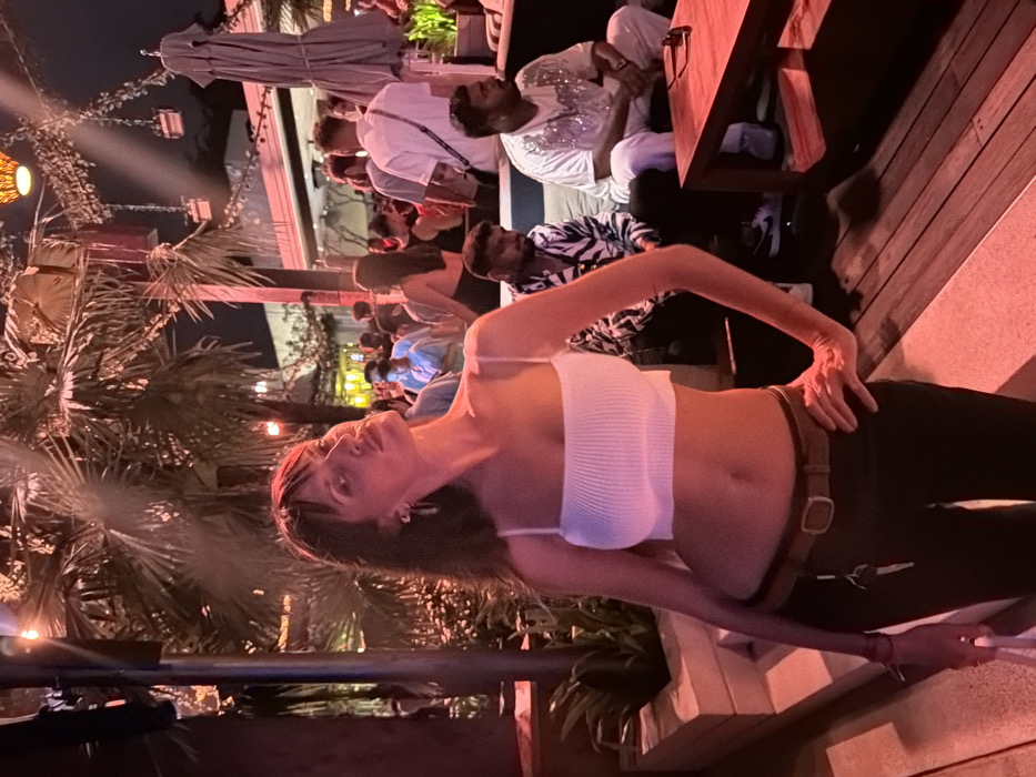 The image depicts an outdoor bar or restaurant scene at night in Pecatu, Indonesia, characterized by warm red and orange lighting. In the foreground, a light-skinned woman with dark hair and bangs stands slightly angled to the right, looking towards the viewer with her head tilted back and chin raised. She is wearing a white ribbed crop top with spaghetti straps, exposing her midriff, and dark pants with a brown belt. A set of keys hangs from her belt on her left hip. Her left arm is by her side, holding a white object, and her right hand rests on her right hip. She has earrings in both ears.
Behind her, the environment features numerous large palm trees illuminated by overhead lights. Several tables and seating areas are visible, occupied by numerous individuals. In the mid-ground, two men are seated on a light-colored sofa. The man on the left wears a black and white zebra-print shirt, while the man on the right wears a white, short-sleeved top with sparkling embellishments. A dark wooden table in front of them has sunglasses resting on it. Further in the background, a crowd of people is visible, some standing and some seated, with various lighting fixtures hanging above. The ground consists of wooden decking and concrete steps. An unbrella is visible on the right.