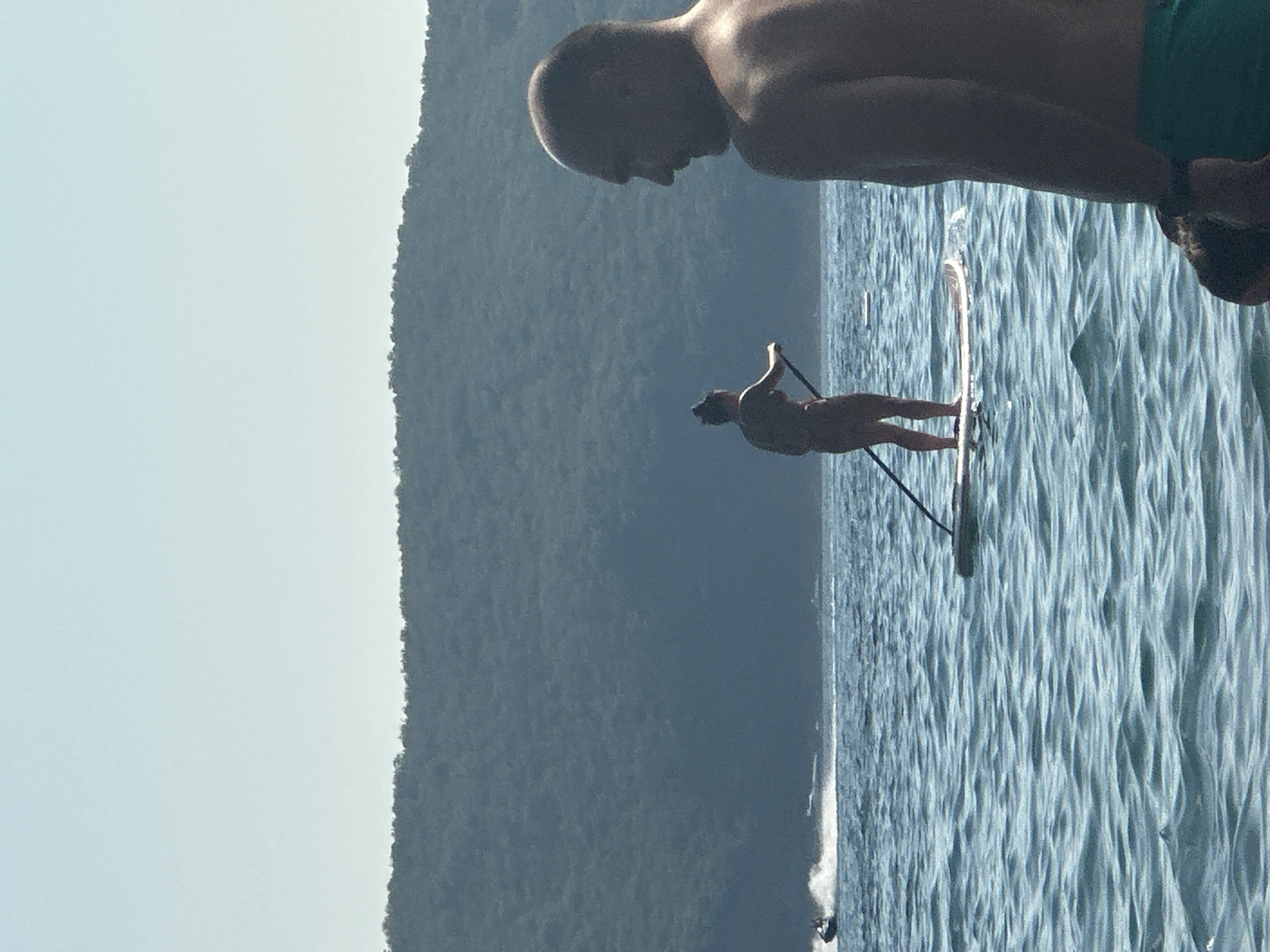 In Boreti, Montenegro, on a body of water, two individuals are engaged in water activities. In the foreground, a man's shoulders and head are visible in profile, facing away from the camera. His skin is bronzed, and he is wearing green shorts. Behind him, a woman is standing on a paddleboard, facing away from the camera and holding a paddle. She is wearing a dark bikini. In the background, a jet ski is moving across the water, creating a spray. Beyond the water, a densely forested hill slopes upwards, with a clear sky overhead. The water is rippled, indicating gentle waves.