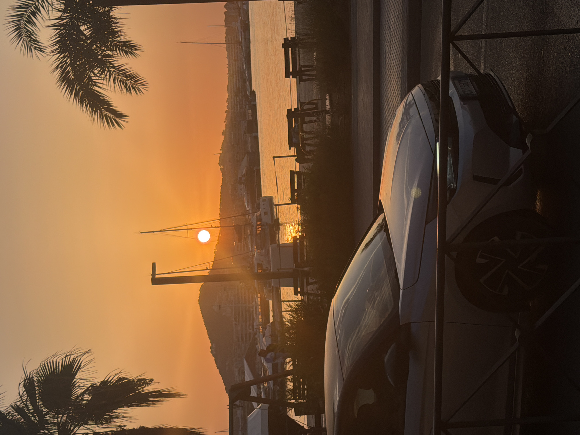 A silver car is parked in the foreground, with its front visible. Behind the car, a waterfront scene unfolds under a dramatic sunset. Palm trees frame the sky, which is ablaze with orange and yellow hues. The sun, a bright orb, is setting over a body of water, likely the Mediterranean Sea, casting a golden reflection. In the background, a hillside dotted with buildings rises, and several sailboats are anchored in the harbor. The location appears to be a coastal area in Ibiza, Spain, possibly near a marina or promenade. The overall atmosphere is serene and picturesque, capturing the beauty of a Mediterranean sunset.
