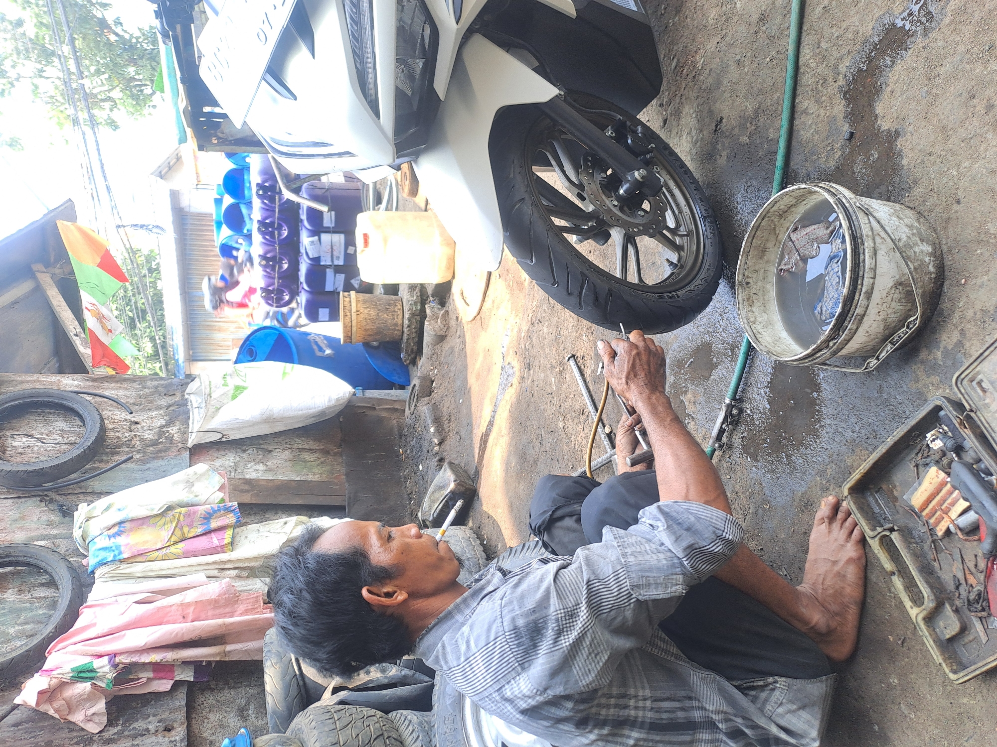 The image depicts an outdoor workshop in Palembang, Indonesia, likely a motorcycle repair shop situated in a somewhat unkempt area. A middle-aged man, appearing to be the mechanic, is crouched down next to the front wheel of a white motorcycle. He is wearing a grey plaid shirt and dark pants. He is holding tools and appears to be working on the tire, possibly checking its pressure or repairing a puncture, as suggested by the presence of a bucket of water and tools. The workshop environment is evident with various objects scattered around, including stacked tires, a toolbox with tools, a white bucket containing water and some items, and several large plastic containers in the background. The ground is dirt and wet in places, indicating recent work or weather. In the background, there are more motorcycles, some drums, and a wooden structure that might be part of the workshop. The lighting suggests it is daytime.