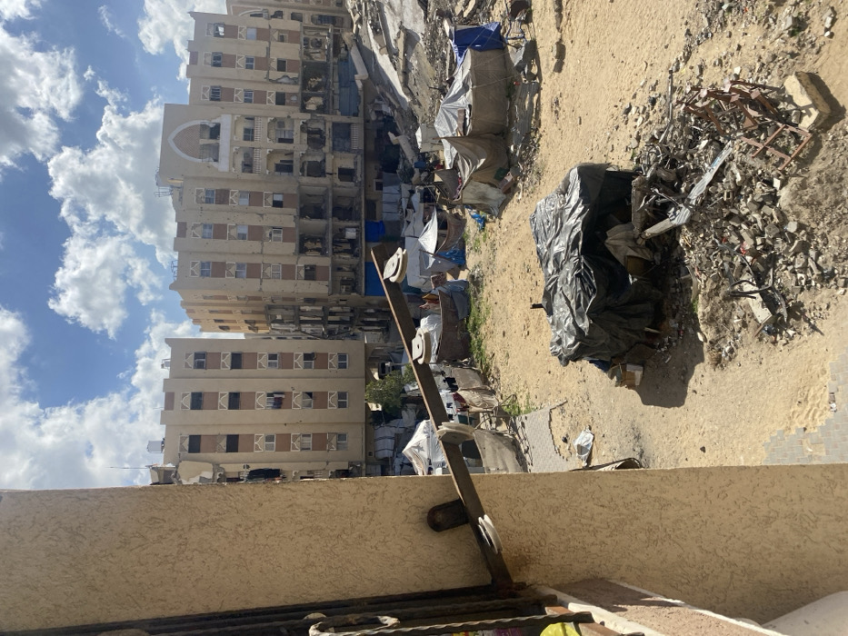 The image depicts an urban landscape in Khan Yunis, Palestinian Territory, characterized by significant destruction and makeshift living conditions under a partly cloudy sky. The photograph is taken from an elevated position, looking out from what appears to be a building or balcony.
In the immediate foreground on the left, a section of a light-colored, stuccoed building wall is visible. Extending horizontally from this wall is a wooden plank, secured by a metal bracket, featuring two white disc-like objects, possibly pulleys or rollers.
The midground consists of a large, open, unpaved area of sandy, disturbed earth. This area is sparsely populated with multiple makeshift shelters constructed from various materials, including tarpaulins (some black, some blue) and light-colored fabric. One prominent, square-shaped shelter covered with a black tarp sits in the lower-center. Adjacent to it, and extending into the lower-right, is a pile of rubble composed of concrete fragments, twisted metal, and miscellaneous debris, with what resembles a twisted metal frame or structure prominently visible. Small trails or paths are discernible between the shelters.
In the background, two large, multi-story buildings dominate the skyline. These structures, approximately six to seven stories tall and light brown in color with decorative architectural elements, exhibit extensive damage. Numerous windows are either shattered or entirely missing, and large sections of their facades, particularly the lower floors, have been destroyed or collapsed, exposing interior rooms and structural elements. The building on the right features a distinctive arched design on its upper floors, also appearing damaged. The overall scene conveys an environment impacted by severe conflict and displacement.