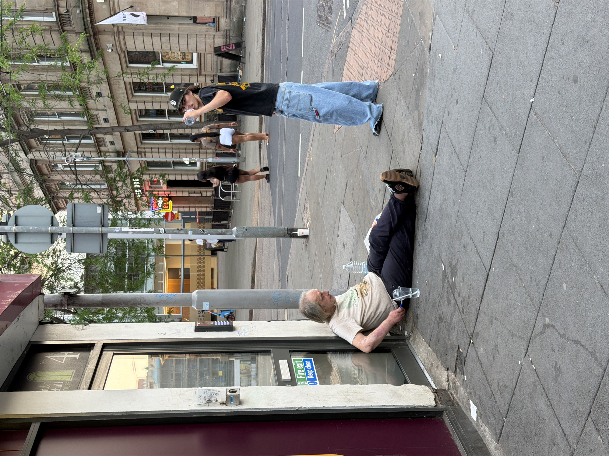 The image shows a street scene in Nottingham, United Kingdom. An elderly man is lying on the pavement next to a shop front with a "Fire Exit, Keep Clear" sign. He is wearing a light-colored t-shirt and dark trousers. A plastic water bottle and a cup are near him on the ground. A person wearing a black t-shirt and very baggy blue jeans is standing on the pavement drinking from a bottle of water. Two other people are walking on the pavement. A traffic light pole is positioned near the shop front. Buildings are visible in the background, including one with a "RED DOG" sign.