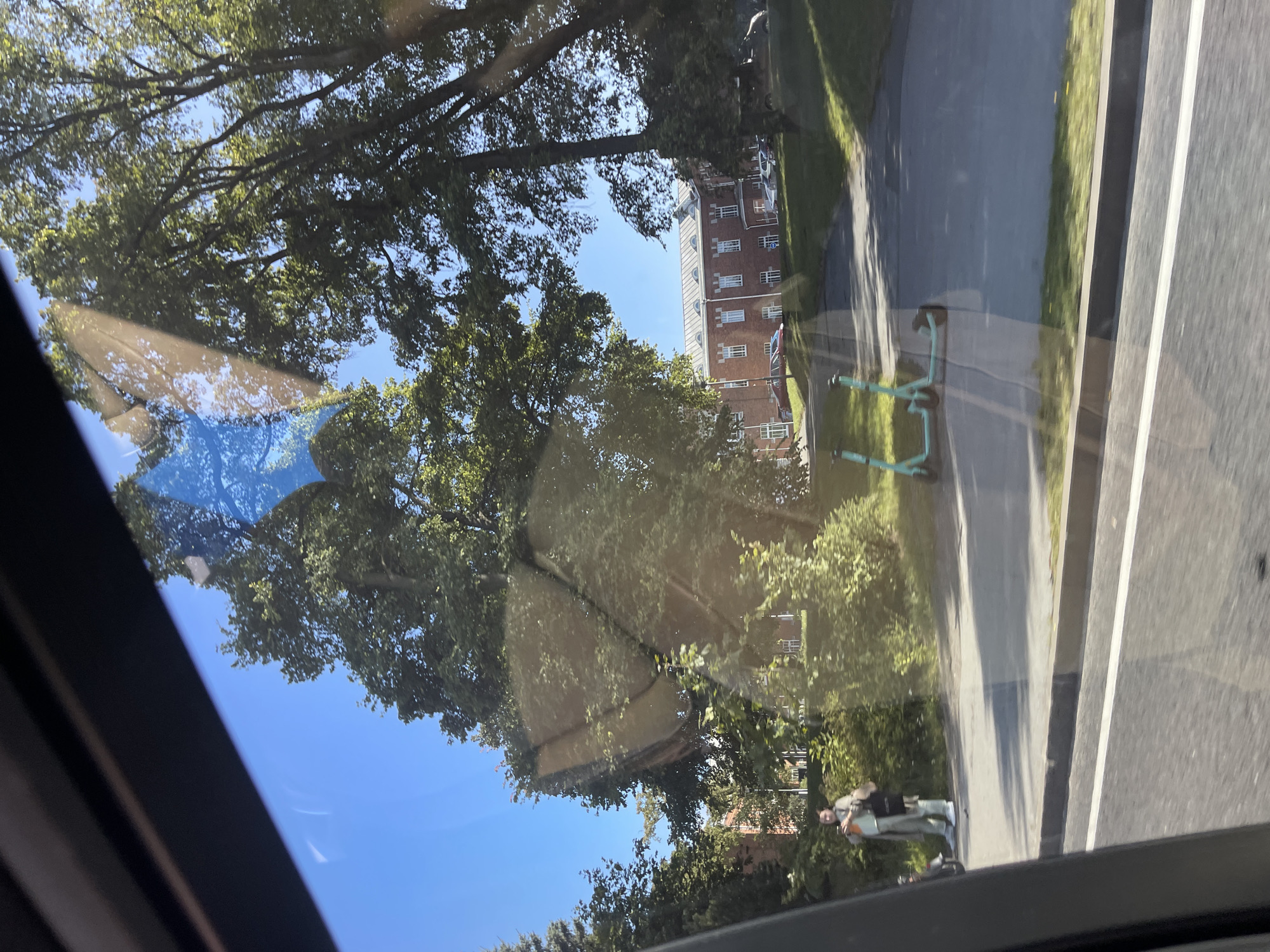 The image depicts a street scene observed through a vehicle window, with reflections on the glass. The setting appears to be a sunny day with a clear blue sky. In the foreground, a paved road with a white line marking is visible. To the right, two light blue electric scooters are parked on the pavement. Further back, a person in light-colored clothing, possibly a trench coat over pants and a shirt, stands holding a bag and a phone. Behind the person, a line of dense green bushes and trees creates a backdrop. In the distance, a large, red brick building with a white roof and numerous windows is situated on a grassy incline. Several cars are parked in front of the building. The overall environment suggests an urban or suburban area, likely in Tallinn, Estonia, given the presence of shared electric scooters common in the city. The image quality is affected by glare and reflections from the window.