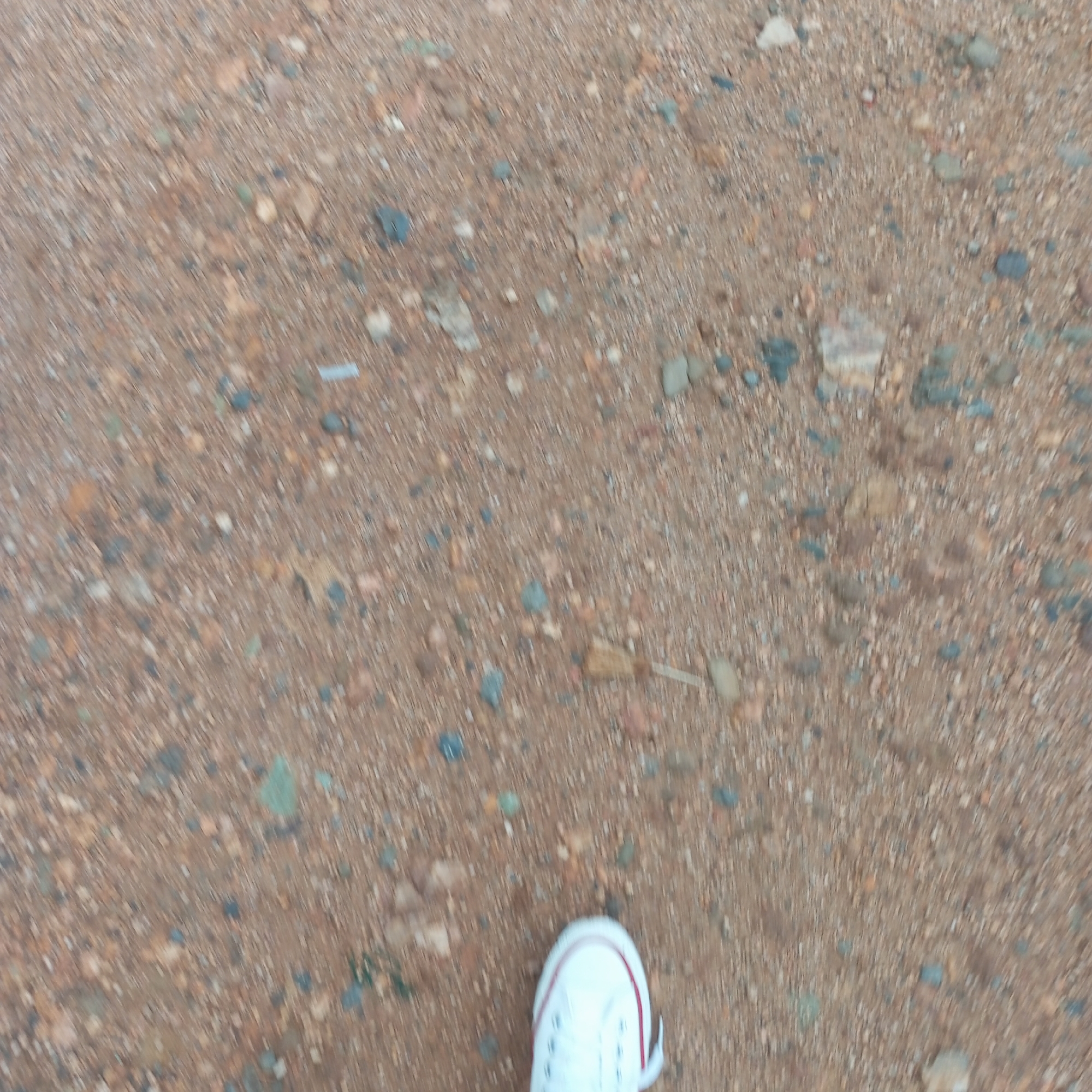 The image depicts a ground surface composed of reddish-brown dirt and small pebbles. A portion of a white tennis shoe with a red stripe is visible in the lower right corner of the frame. The image appears to be taken from a first-person perspective, looking down at the ground. There are small rocks in the dirt. It can not be determined that the location is in Mahiakalo ward, Kenya, as there is no evidence of that location in the image.