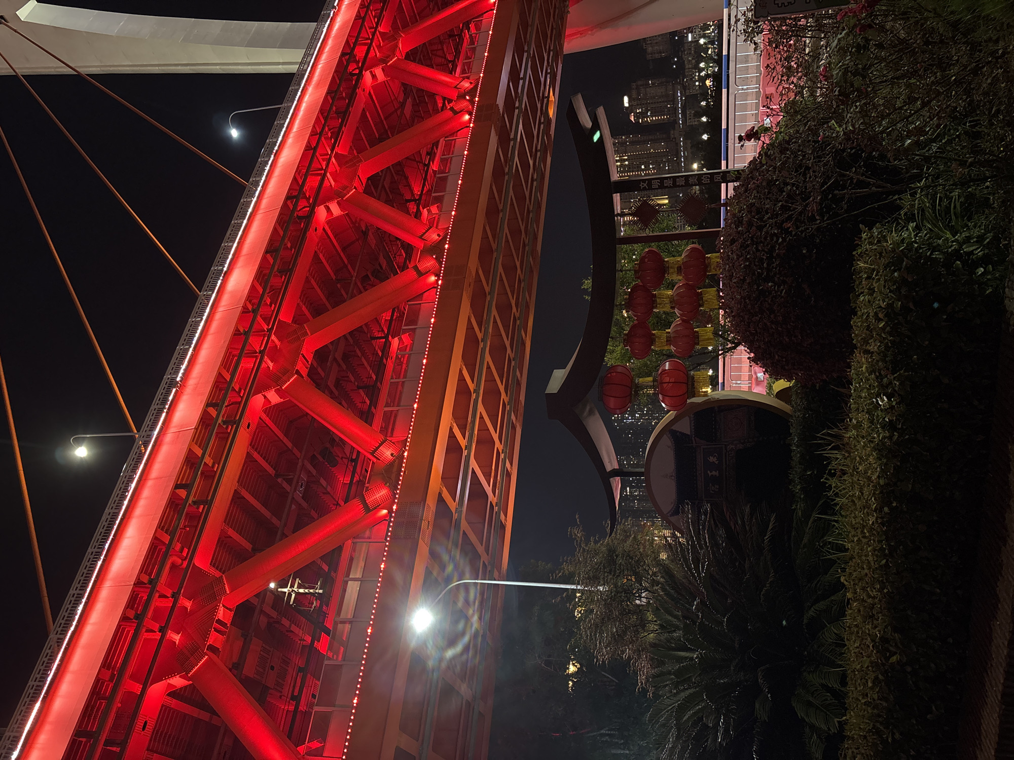 The image depicts a section of the illuminated Qianximen Bridge in Yuzhong District, Chongqing, China, at night. The bridge's red steel structure is prominently featured. Adjacent to the bridge are decorative elements: vertical rows of red lanterns and greenery, suggesting a park or public space alongside the river. In the background, the city skyline is visible. Streetlights illuminate the area.