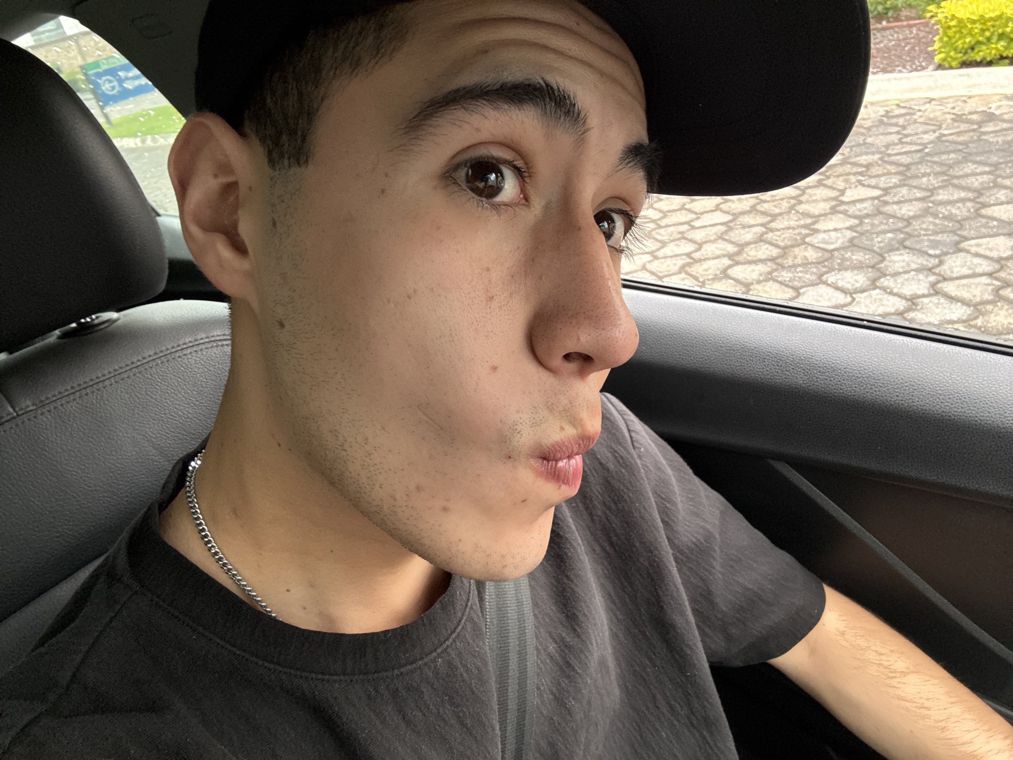 The image captures a young male situated in the passenger seat of a vehicle, in what is identified as San Bernardino Tlaxcalancingo, Mexico. The individual, with light to medium complexion, dark hair, and visible facial stubble, wears a dark t-shirt and a silver-toned cuban link chain. A dark baseball cap is partially visible on his head. His dark brown eyes are wide and directed towards the viewer, while his lips are pursed into a distinct pouting or kissing expression. A seatbelt is diagonally secured across his chest. The car's interior features dark grey leather seating and dark grey plastic door trim. Through the vehicle's windows, exterior elements are visible: on the left, behind a dark headrest, a distant, partially obscured green and blue sign, possibly indicating "Park" or "Parking," is discernible amidst green foliage and a damp-appearing road surface. On the right, a textured ground of interlocking light-colored pavers or cobblestones is brightly lit, contrasting with a vibrant yellow-green bush beyond it.