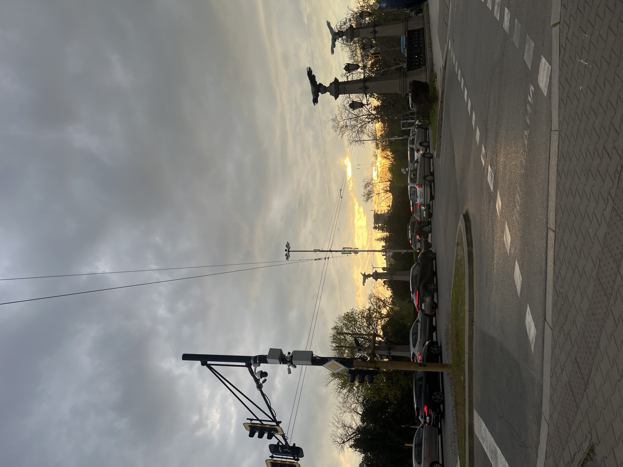 The image presents an urban street scene captured during sunset or golden hour, looking towards the horizon where the sun's warm, golden light bursts through a heavily clouded sky. This dramatic illumination casts a soft, hazy glow across the upper part of the scene and silhouettes many of the foreground and mid-ground elements.
In the mid-ground, two prominent bronze statues of eagles, mounted on ornate stone pedestals, are distinctly silhouetted against the bright, low sun. These statues, along with visible streetlights on their pedestals, are characteristic of Eagles' Bridge (Orlov Most) in Sofia, Bulgaria. Behind the bridge structures, bare trees, suggesting late autumn or early spring, line the road, with some distant buildings vaguely visible.
The foreground features a multi-lane road with white dashed lines marking a lane or parking area, and a solid white line separating it from a wide sidewalk on the right. The sidewalk is paved with gray, interlocking patterned stones. Numerous vehicles, predominantly dark-colored sedans and SUVs, are parked along the road, some with illuminated taillights, indicating either stationary traffic or parked cars.
To the left, a tall pole supports a complex array of traffic lights, electronic sensors, and possibly surveillance cameras, with a yellow diamond-shaped sign mounted nearby. Overhead utility or trolley wires stretch across the sky. One traffic light displays a green arrow, and another appears yellow. The sky is largely overcast with scattered clouds, creating a dynamic backdrop for the setting sun. No discernible text is visible within the image.