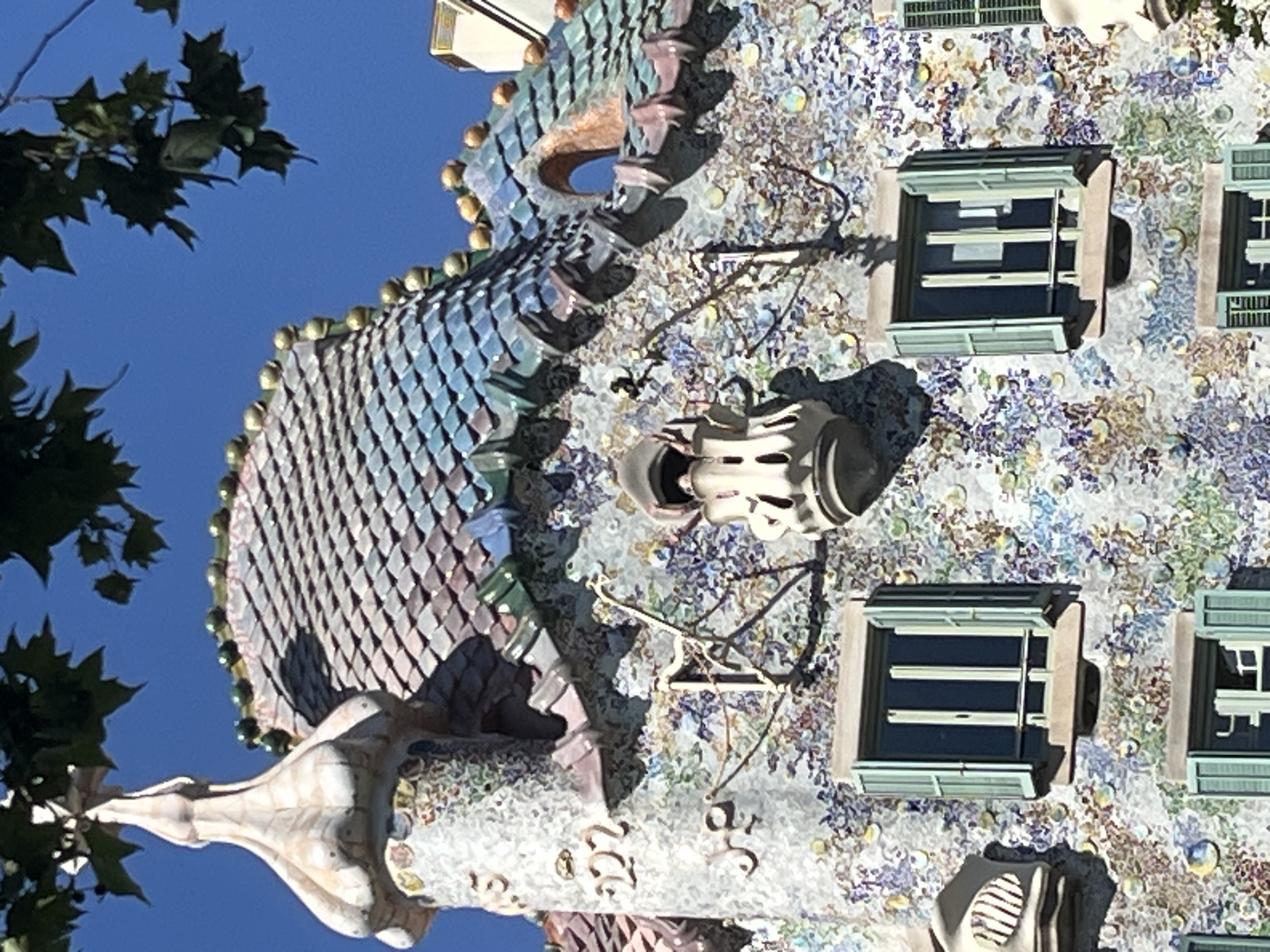 The image shows a close-up view of the facade of Casa Batlló, a renowned building located at Passeig de Gràcia in Barcelona, Spain. The facade is characterized by its organic, undulating design and a vibrant mosaic of colorful ceramic tiles and glass fragments.
The upper part of the building features a roof with a shimmering, scale-like pattern in shades of blue, green, and purple, reminiscent of a dragon's back, topped with golden spheres. A distinctive turret, shaped like a mushroom or a fantastical spire, rises from the left side of the roof, adorned with delicate carvings and embellishments.
Below the roofline, the wall is covered in a complex mosaic of small, irregularly shaped pieces of ceramic and glass, creating a fluid, organic texture with floral and abstract patterns. This mosaic incorporates various colors, including blues, purples, greens, yellows, and white, with reflective elements that catch the sunlight.
Several windows are visible. The most prominent are two rectangular windows with light green shutters that are partially open, revealing dark window panes. These windows have a projecting shelf below them. Above the left window, a sculpted element resembling a balcony or decorative outgrowth extends from the wall. Further up, a more elaborate, skeletal-like structure with a small opening is visible, also adorned with the mosaic.
The image is taken on a clear, sunny day, with a bright blue sky visible in the background. Green leaves from a tree partially overhang the top of the building, casting dappled shadows. The overall impression is one of architectural artistry and vibrant, whimsical design, showcasing Antoni Gaudí's unique style.