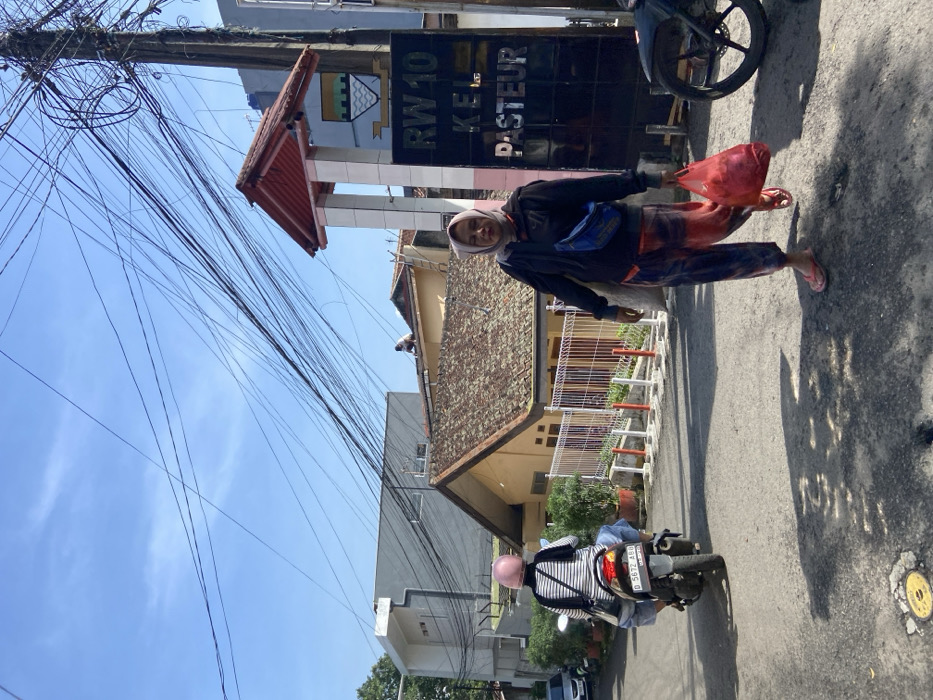 The image depicts a street scene in the Pasteur neighborhood of Bandung, Indonesia, located at coordinates -6.892 latitude and 107.599 longitude. A woman wearing a headscarf and carrying a red bag walks down the street towards a sign that reads "RW 10 KEL PASTEUR." A motorcycle with a pink helmet on the seat is parked to the left of the sign. The sky is clear blue with a few scattered clouds. There are numerous power lines extending from a utility pole in the background. There are two buildings in the background with tiled roofs. The street is paved with asphalt and has a slight incline. The scene is a typical street in a residential neighborhood.