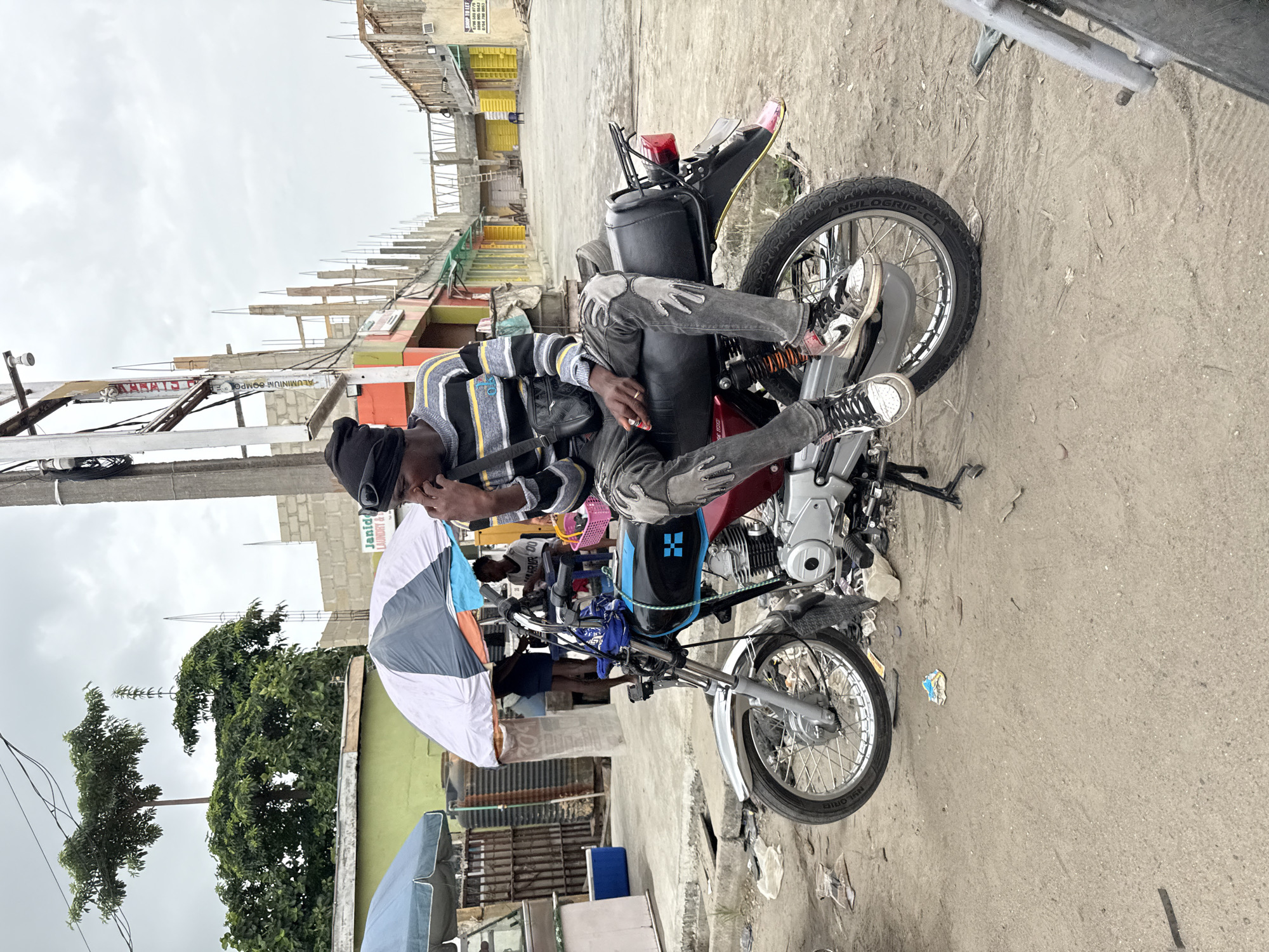 An individual is sitting on a motorcycle in what appears to be a street in Ikorodu, Nigeria. The motorcycle is parked on a concrete surface next to buildings and structures, some of which appear to be under construction. The individual is wearing a striped sweater, dark pants, and shoes, and seems to be using a mobile phone. An umbrella is also visible near the motorcycle.