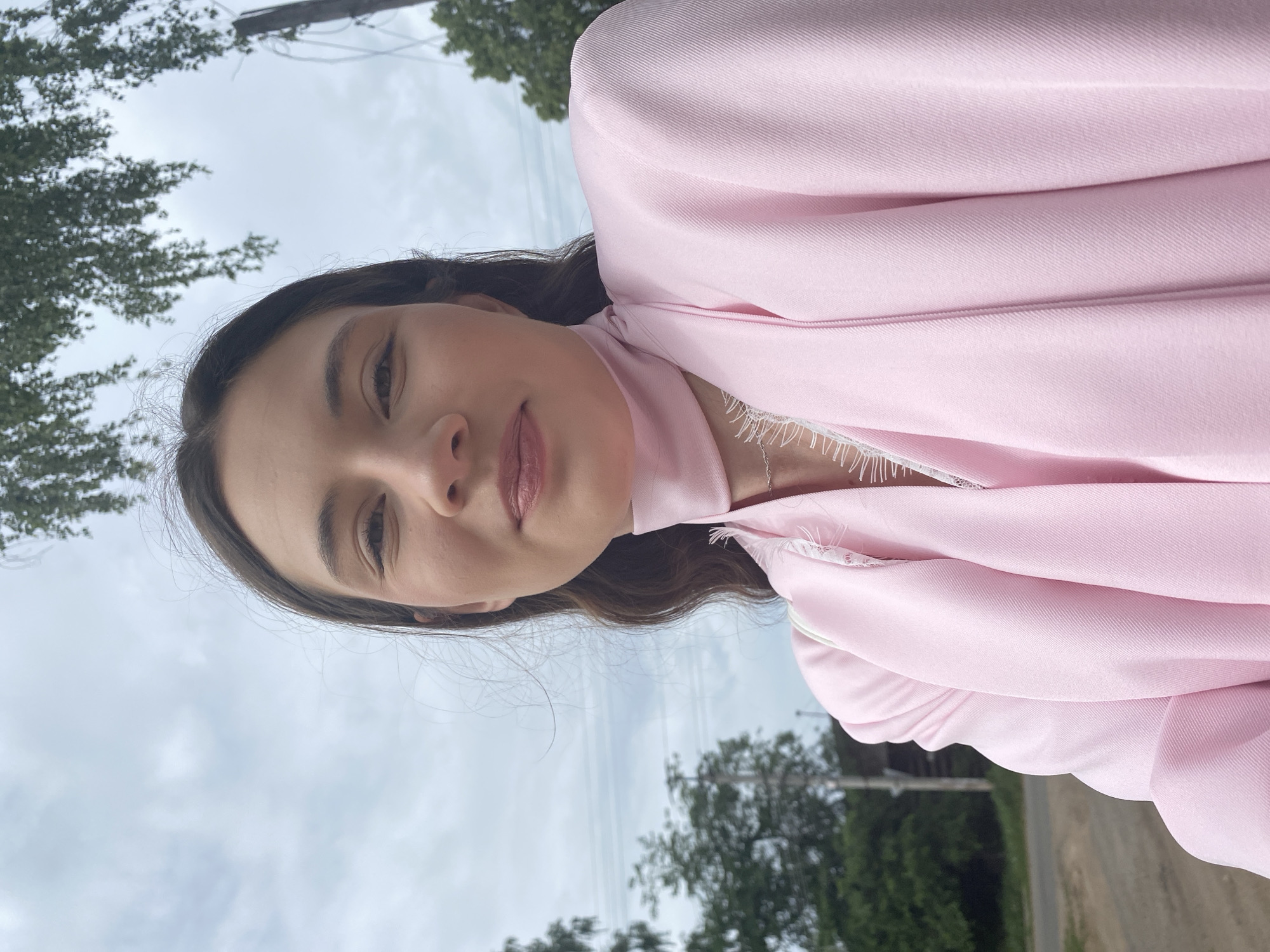 A young woman is centered in a selfie taken outdoors. She has brown hair, and is wearing a pink blazer with a scarf, with white lace detailing around the neckline. The sky is overcast, and power lines are visible in the background. Green foliage and trees appear along the upper and lower edges, suggesting an outdoor setting, possibly near a road. It is unknown if the location is in Votkinsk, Russia.