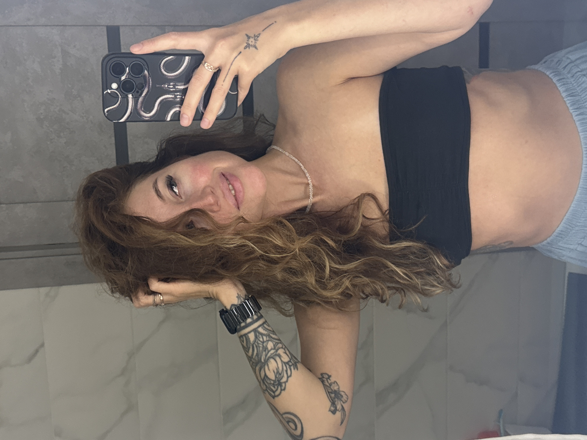 A woman with long, curly, reddish-brown hair, wearing a black strapless top and light blue pants, is taking a selfie in a bathroom. She has tattoos on her left arm and a small tattoo on the back of her right hand. She is also wearing a beaded necklace and rings on her fingers. The bathroom appears to have marble-patterned tiles on the walls and a gray countertop. The overall scene suggests a casual, personal moment captured in a home environment. The location is not discernible from the image itself.