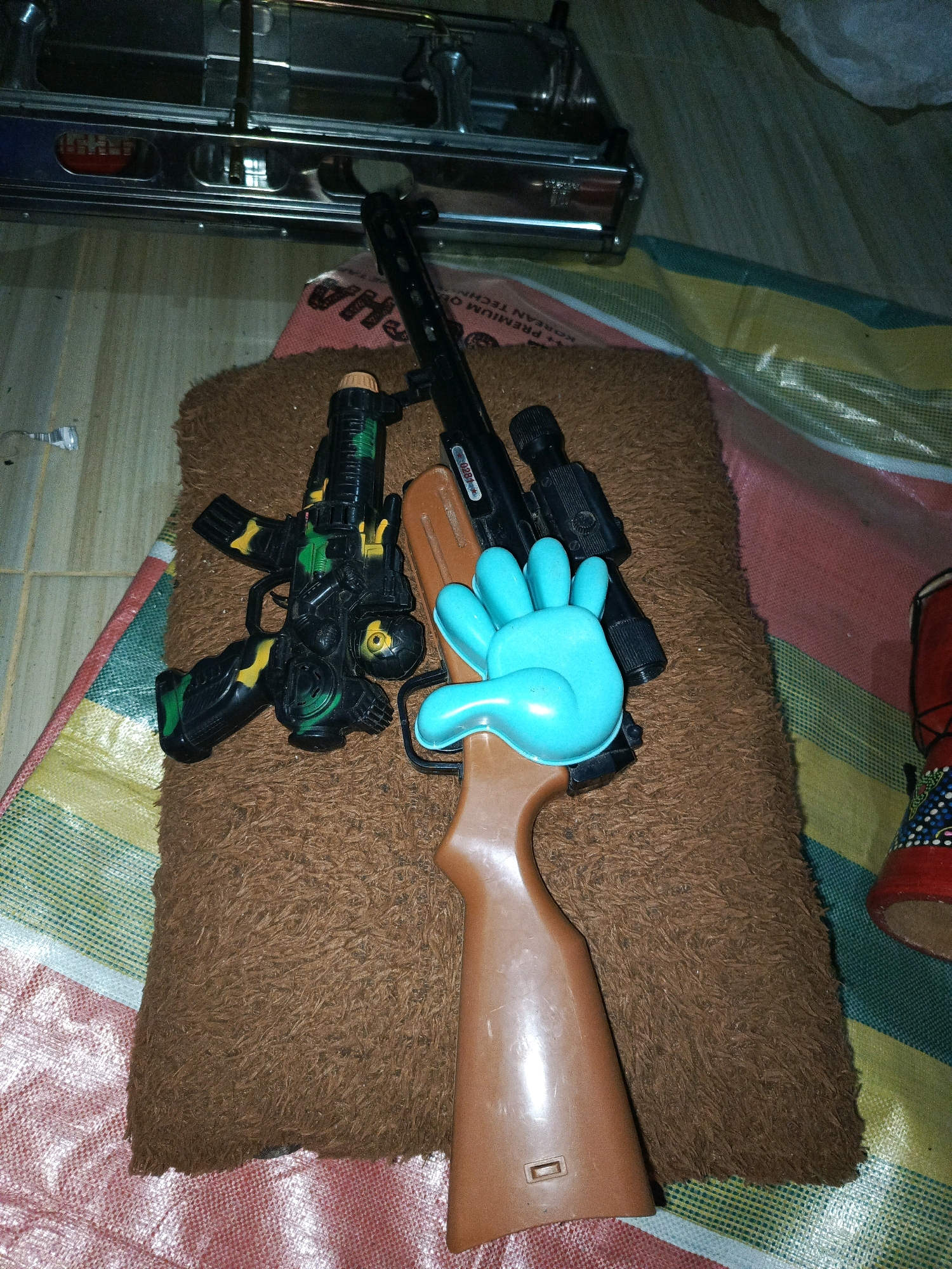 The image shows a collection of toy guns arranged on a brown fuzzy mat. The mat is placed on top of a colorful striped fabric. In the background, a metal object, possibly a piece of furniture or equipment, is visible. One of the toy guns is brown and resembles a rifle, with a bright blue plastic hand placed on its stock. Another toy gun is black and green with camouflage detailing. A third, smaller black toy gun is also visible. The overall scene suggests a casual indoor environment. The image does not provide any specific information about the city of Megang Sakti I, Indonesia.