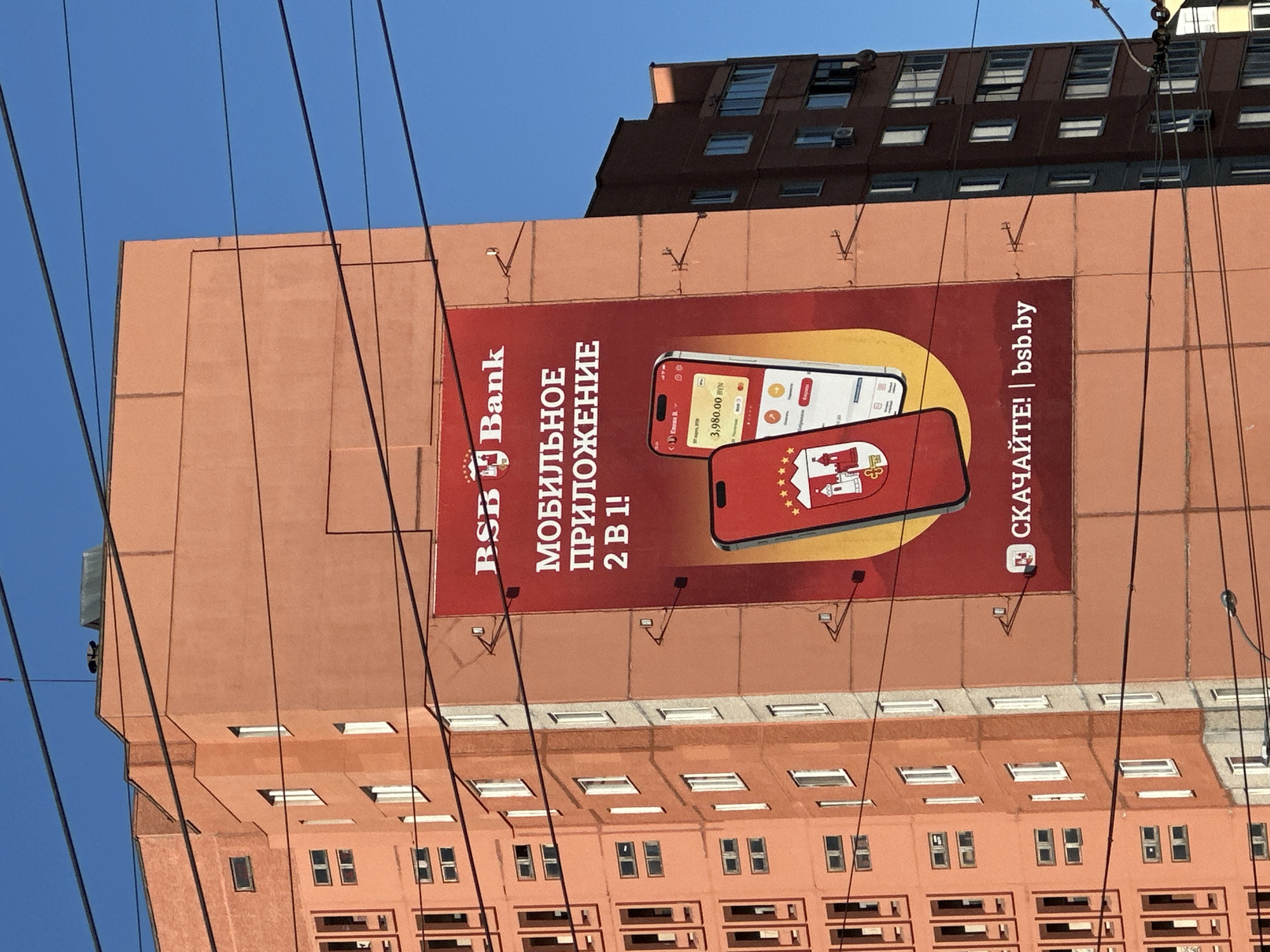 The image shows a large advertisement for BSB Bank on the side of a building. The advertisement features two smartphones displaying the bank's mobile application, and includes the text "MOBILE APPLICATION 2 IN 1!" and "DOWNLOAD!". The building is a multi-story structure with numerous windows. The sky is clear blue. It is a building located in the city of Minsk, Belarus.