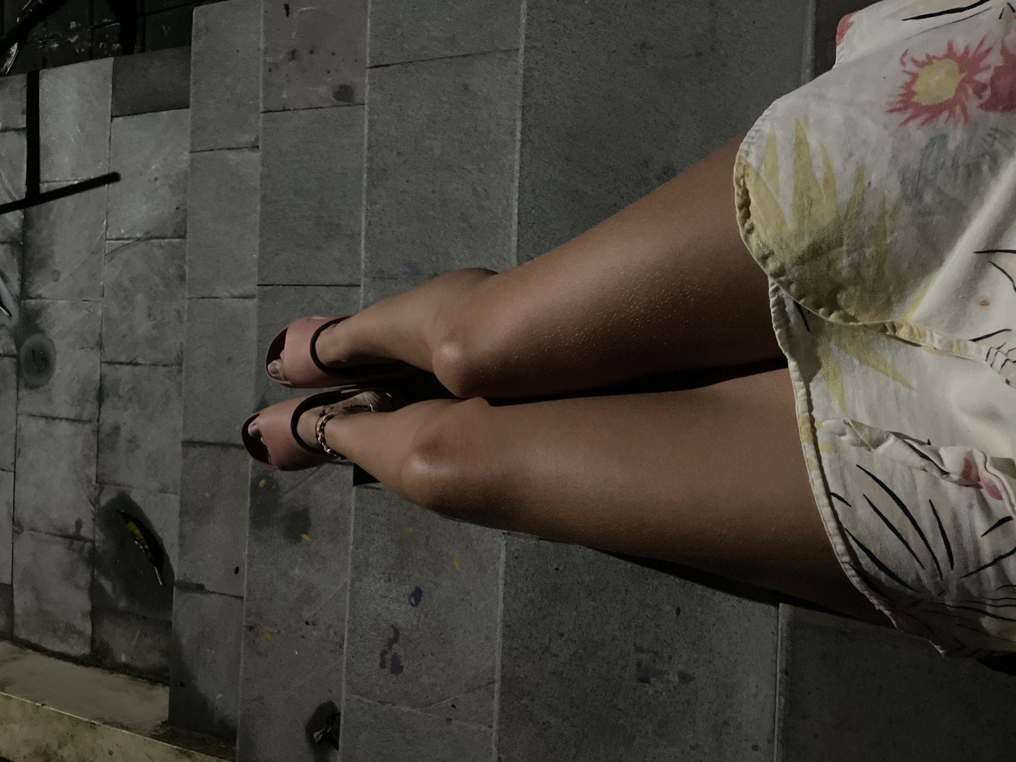 The image captures a person's legs and lower torso, dressed in a patterned garment, resting on a series of stone steps. The scene is dimly lit, suggesting nighttime or a low-light environment.
The subject is a person, visible from just above the knees downwards, with their legs extended. They are wearing light pink, open-toed sandals and a beaded bracelet on their right ankle. The skin on their legs appears tanned. Their lower torso is covered by a light-colored dress or skirt featuring a pattern of pink and yellow floral or leafy motifs, interspersed with black linear designs.
The setting consists of grey stone steps, which appear to be constructed from large, rectangular blocks. The texture of the stone is visible, and some steps show faint specks of color, possibly paint or debris. A small, light-colored butterfly or moth is present on one of the lower steps to the left.
The activity appears to be resting or posing on the steps. The low light creates a subdued atmosphere, emphasizing shadows and the textures of the skin, fabric, and stone. There is no visible text in the image. The location context is Kuta Selatan, Indonesia.