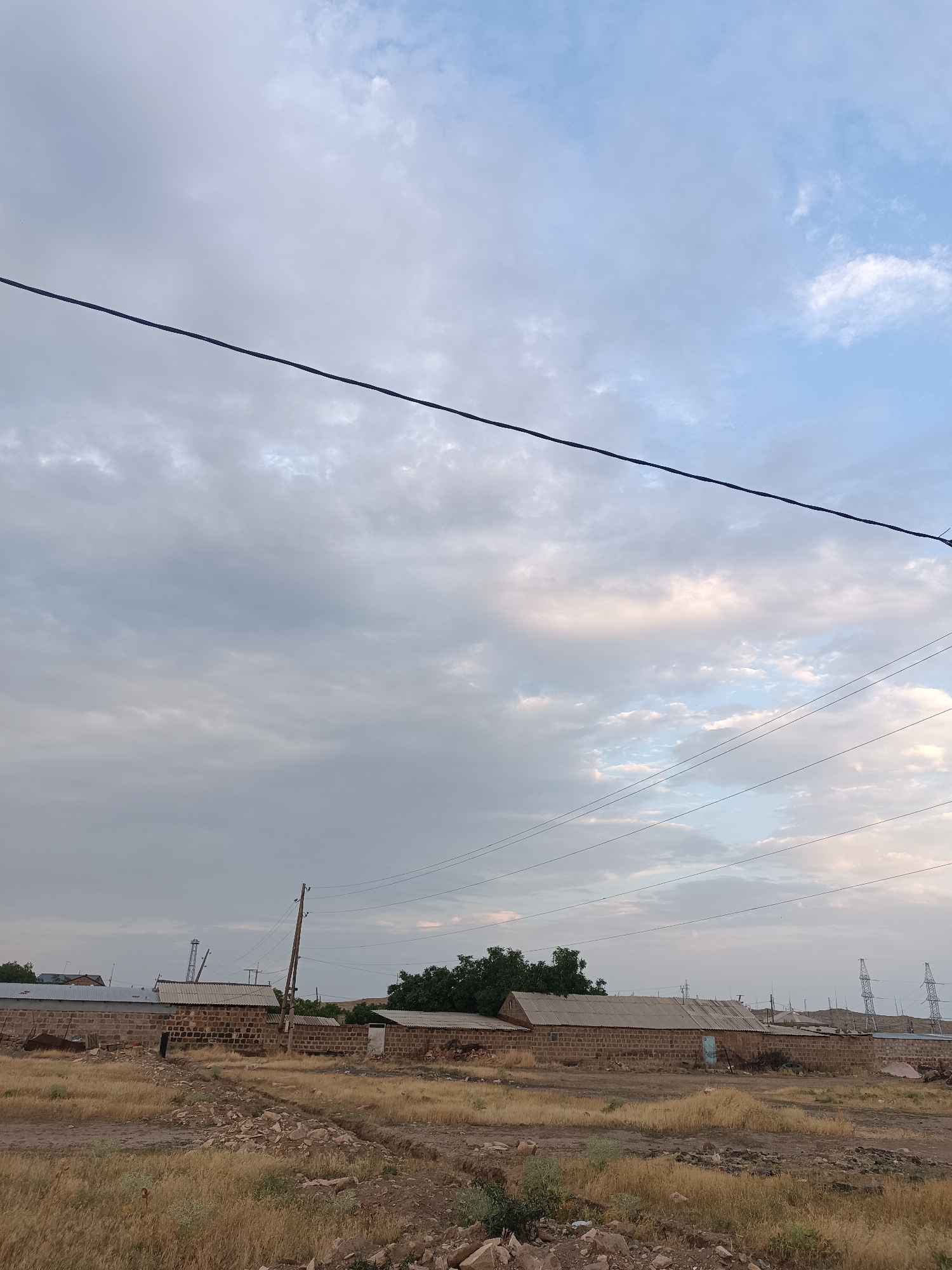 This is a horizontal, eye-level, medium-shot photograph taken in the city of Talin, Armenia. The scene depicts a collection of modest, single-story buildings constructed with reddish-brown brick walls and corrugated metal roofs. The structures appear to be low-rise, likely residential or utility buildings, with some showing signs of wear. A thick, dark electrical wire runs diagonally across the upper portion of the image, from the top left to the bottom right. Another set of thinner wires is visible lower down, strung between wooden poles.
The ground in front of the buildings is covered with dry, yellowed grass and sparse patches of bare earth and small rocks. A dirt path or track is visible in the foreground. In the background, several tall, lattice-like electrical pylons stand against a partly cloudy sky. The sky itself transitions from a lighter blue near the horizon to a softer, overcast grey with hints of pink and white clouds overhead. The overall impression is one of a rural or semi-rural setting, possibly on the outskirts of the city, characterized by functional, utilitarian architecture and a dry, arid landscape. The lighting suggests it is either early morning or late afternoon. There are no people or animals visible in the frame.