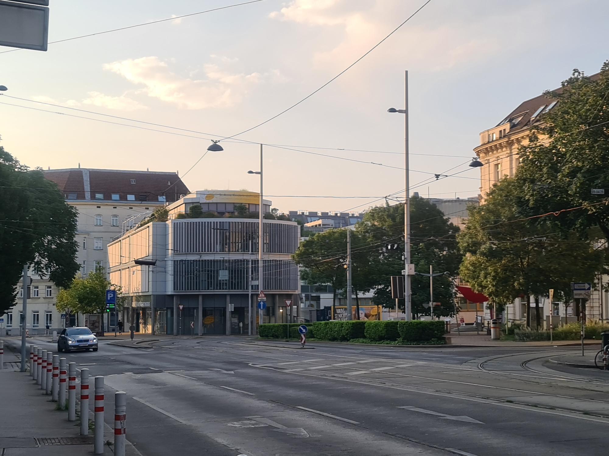 The image depicts a street intersection in Vienna, Austria, during a warm, late afternoon or early evening. The sky is a pale orange and yellow, with some wispy clouds. Overhead power lines crisscross the scene. On the left, a silver sedan is driving towards the viewer. A sidewalk with bollards separates the pedestrian area from the street.
Several buildings are visible. To the left, a light-colored building with a red-tiled roof and multiple windows dominates the foreground. Adjacent to it, a modern building with a glass facade and vertical metal louvers stands out. Its top floor features a green, landscaped terrace. Further back, other buildings with various architectural styles are visible, including a yellow building with arched windows and a dark-tiled roof.
On the right side of the intersection, a tree-lined area with dense green bushes separates the road from more buildings. One of these buildings has a mansard roof and several windows. A large advertisement, partially obscured, is visible on a blue structure in the background.
Street furniture includes traffic lights, street lamps on tall poles, traffic signs (including a blue sign with a white arrow indicating a turn, a triangular yield sign, and a blue parking sign), and tram tracks embedded in the asphalt. A bicycle is parked on the right side of the street near the curb. The road itself is marked with white lines, indicating lanes and pedestrian crossings. There are at least two people visible: one walking on the sidewalk in front of the modern building and another, possibly a pedestrian crossing the street, further in the background. The overall impression is of a well-maintained urban environment on a clear day, with the characteristic infrastructure of a European city.