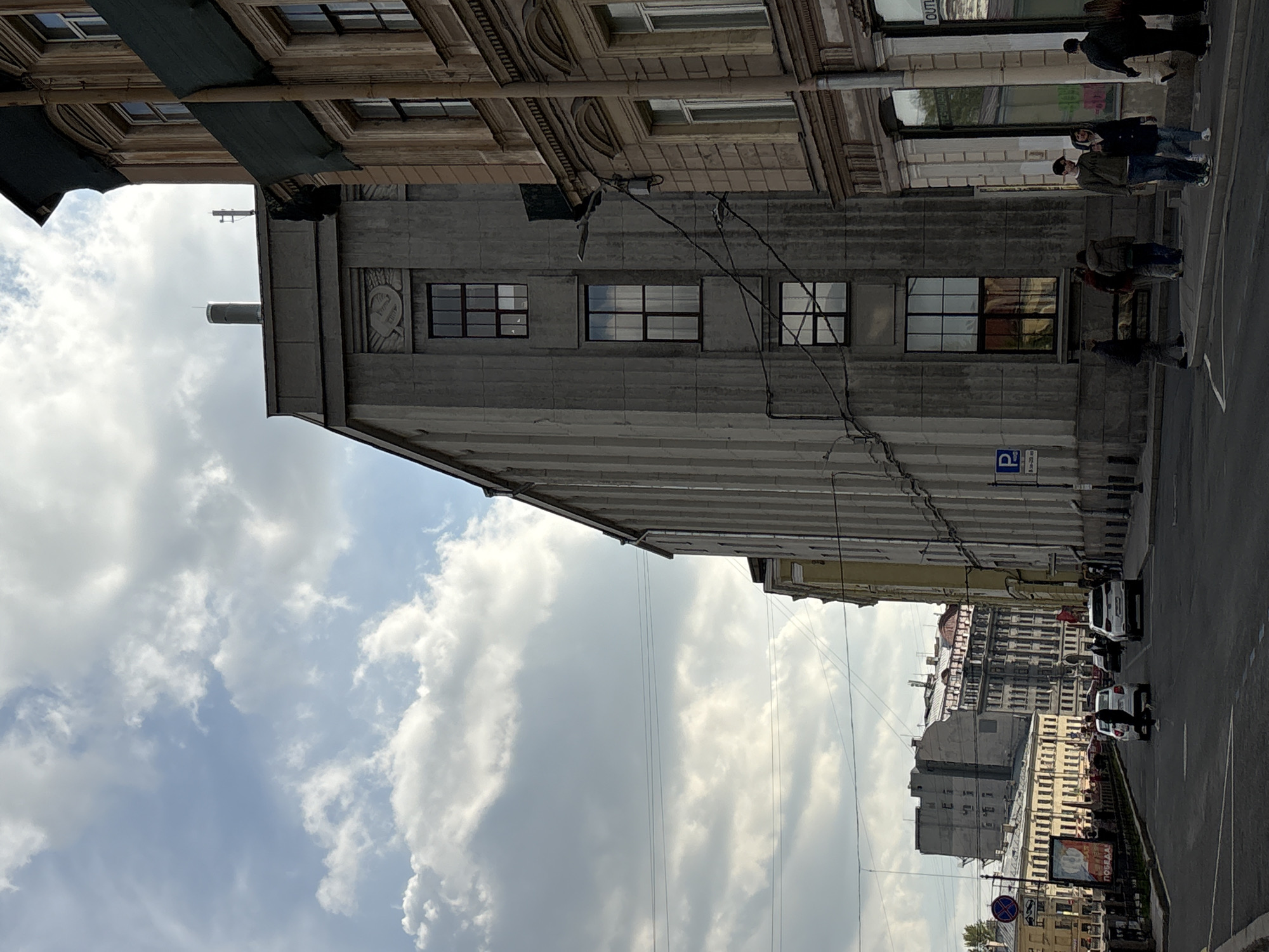 Captured at an upward angle, the photograph presents a city street in Saint Petersburg, Russia. The sky, a mix of blue and white clouds, serves as a backdrop to a gray building featuring rows of windows and vertical architectural lines. A metal pipe extends from the building’s roof.
A portion of an older building with a more ornate facade is visible above the gray structure, with dark awnings extending over its windows. Power lines crisscross in front of the buildings.
On the right side of the frame, pedestrians walk along the sidewalk adjacent to a street with parked and moving vehicles. The buildings beyond the street create a layered urban environment. A parking sign with the letter "P" is affixed to the gray building.