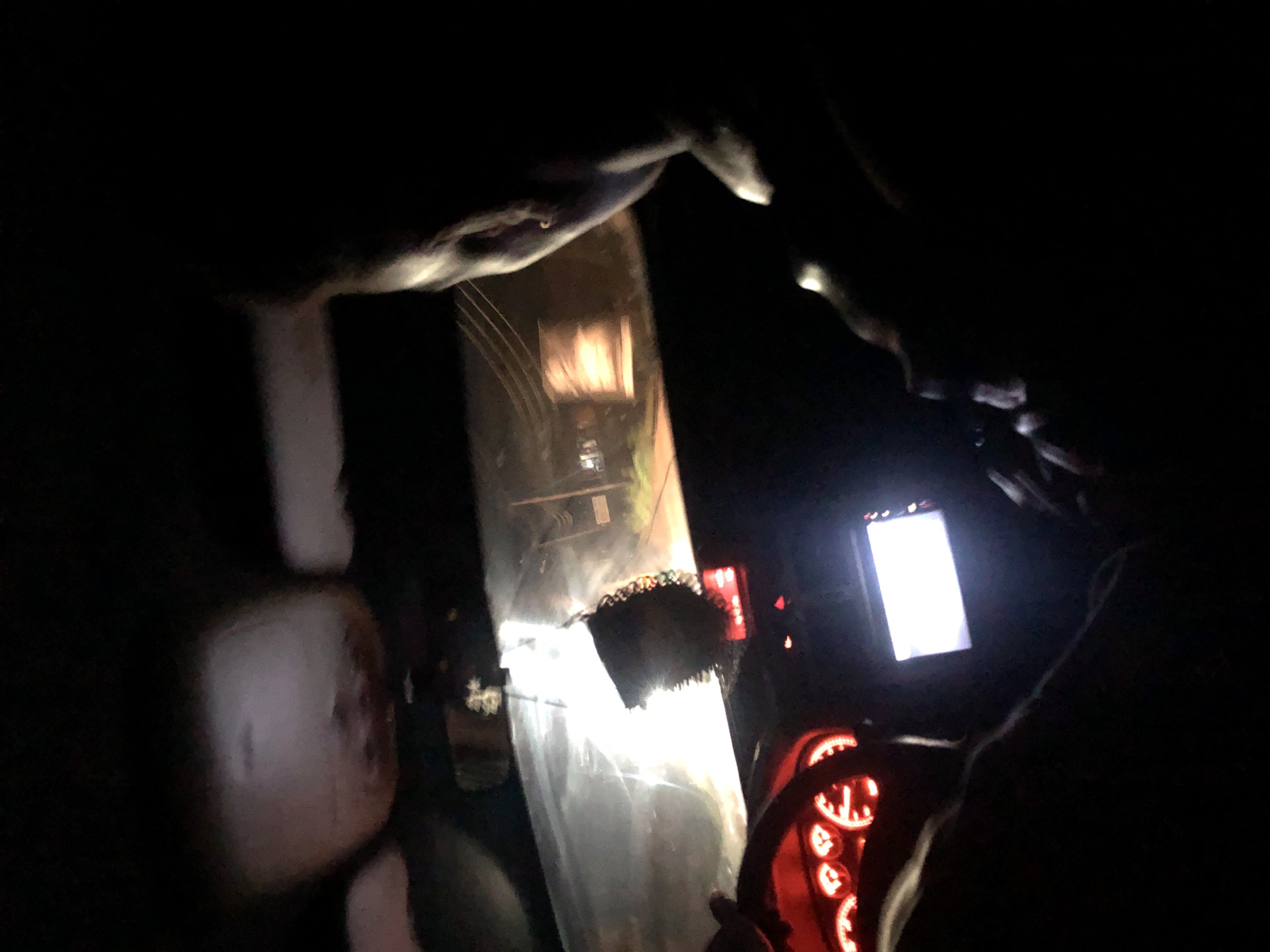 The image depicts the interior of a car at night. A person's head and shoulder are visible on the right side, silhouetted against the darkness. In the foreground, on the left, a glowing red dashboard with illuminated gauges is visible, indicating the car is operational. A car stereo or display screen is also visible in the center, emitting a bright white light. Outside the windshield, the scene is illuminated by streetlights and the headlights of the vehicle, revealing a road and some structures. A hanging air freshener is visible from the rearview mirror. The overall impression is of a night drive through a city, possibly Rukuba Road, Nigeria.