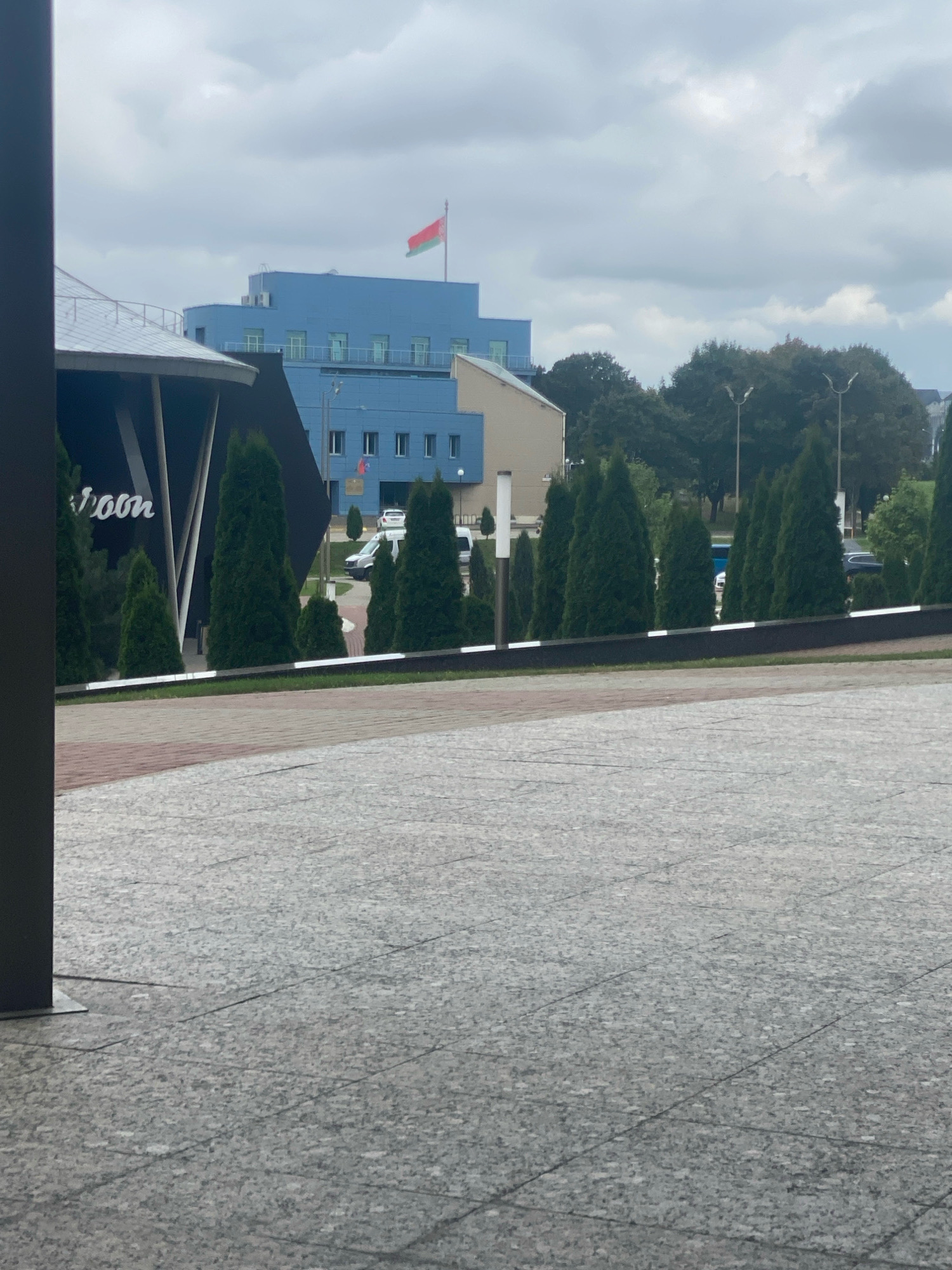 The image shows a view of a government building in Minsk, Belarus. The building is light blue with a red and green flag on top, which is the flag of Belarus. There are trees in front of the building, and a paved area in the foreground. The sky is cloudy. The building appears to be modern, with many windows. The paved area is made of large, grey stones. The trees are tall and thin, and appear to be evergreens. There is also a dark, modern structure on the left side of the image, partially obscuring the view of the building and trees.