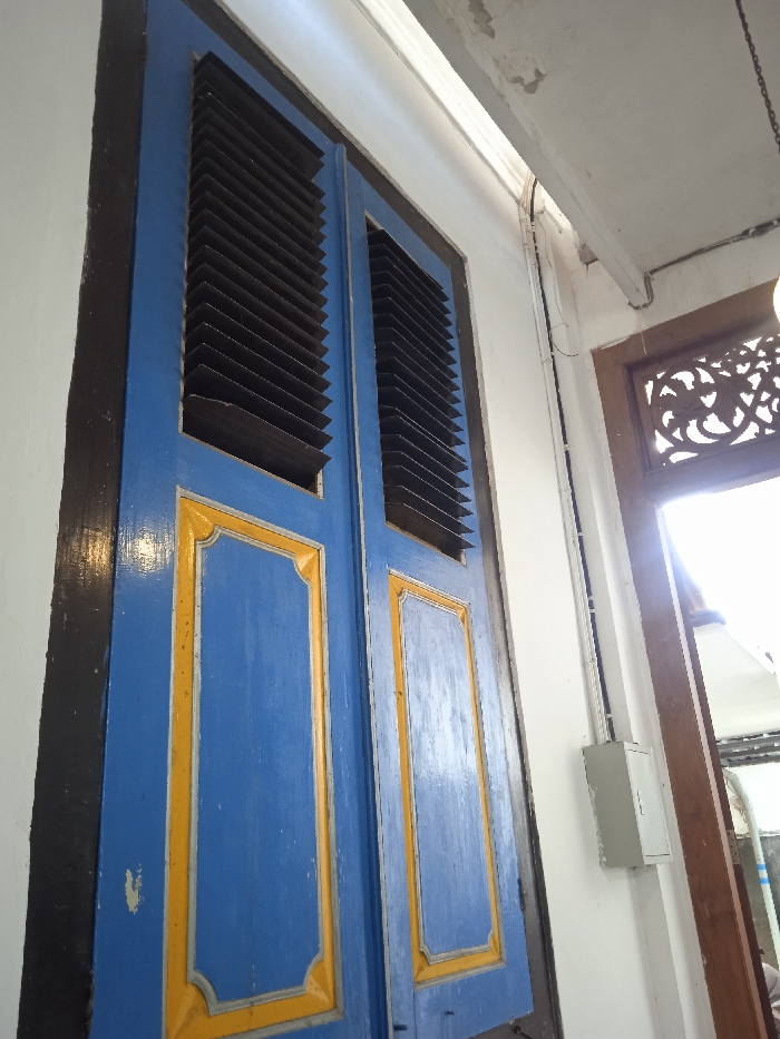 The image shows a pair of blue wooden doors with yellow trim and black frames. The doors are closed and have a set of black louvered panels on the top half. The doors are located in a hallway with white walls. The hallway is brightly lit with a white ceiling and a wooden doorway leading to another room. The wooden doorway has ornate carvings, and a bright light is visible through the doorway. There are also electrical wires running down the wall next to the doors. The location of the image is on Jalan Slamet Riyadi, Surakarta, Indonesia (latitude: -7.559, longitude: 110.783).