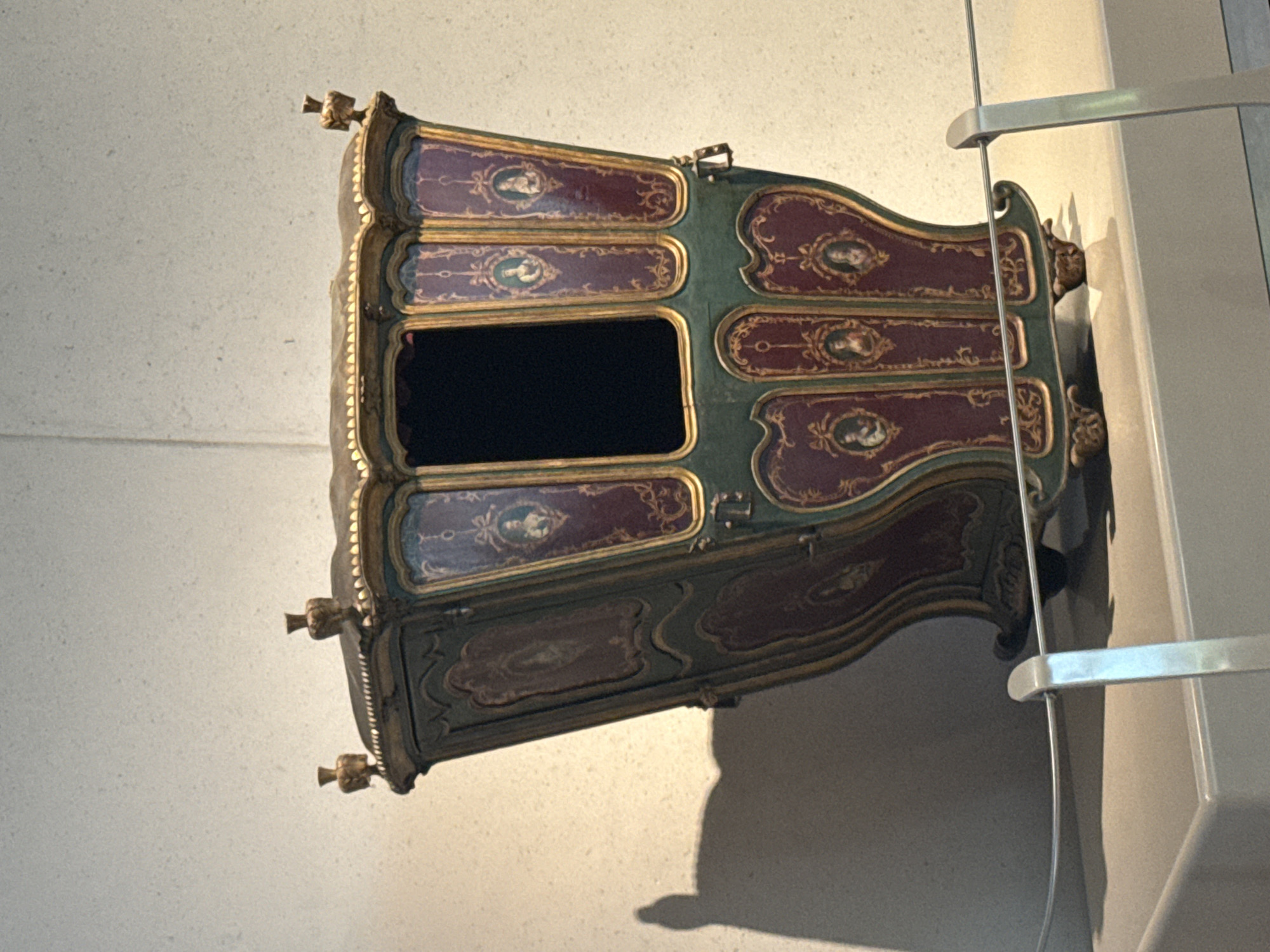 The image shows a decorative, antique sedan chair displayed against a wall. The chair is predominantly maroon and green with gold trim and features painted portraits within decorative frames. It has a dark, rectangular opening where the occupant would sit. The sedan chair is positioned near a metal railing or barrier. The location is not evident from the image, so San Bernardino Tlaxcalancingo, Mexico, cannot be confirmed.
