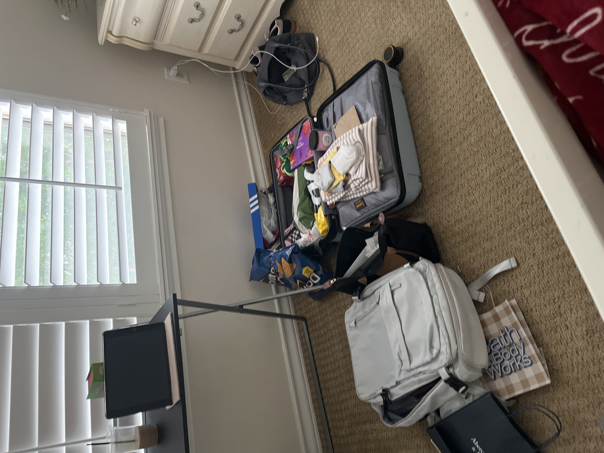 The scene is indoors, likely a bedroom, cluttered with luggage and travel-related items. An open suitcase lies on the carpeted floor, filled with clothes and various personal belongings. Next to it, there is a white backpack and shopping bags. A black backpack is also present. A desk with a tablet is positioned near a window with blinds. A dresser is partially visible in the upper left corner of the frame. The location appears to be a private residence. There is no evidence to suggest it is in San Antonio, United States of America.