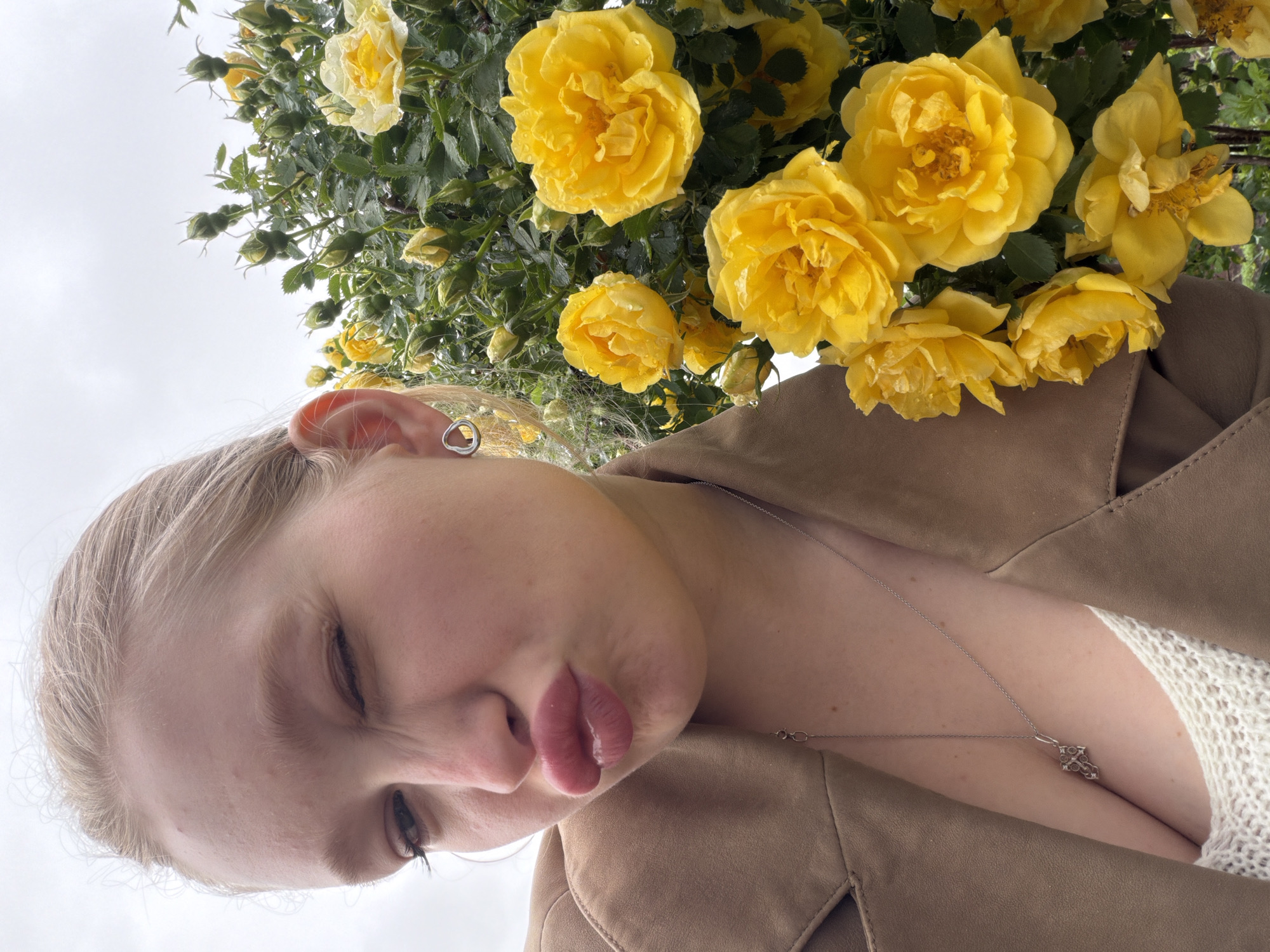 A close-up, slightly angled photograph shows a person with light hair and skin, positioned against a backdrop of yellow flowers. The person is wearing a brown jacket over a white, textured top. A silver necklace with a pendant is visible. The flowers are predominantly yellow roses, some with water droplets on the petals. The background appears to be an overcast sky, indicating outdoor setting. The location is not discernible from the image alone.
