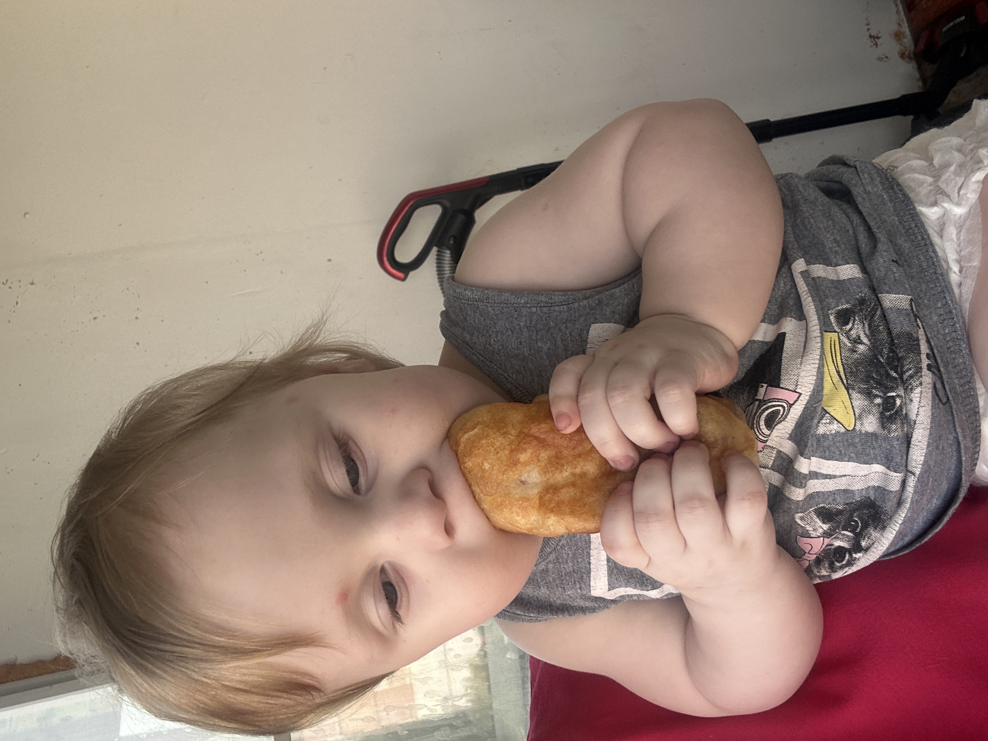 A young child with blonde hair and a red mark on their forehead is sitting and eating a piece of food, likely bread or pastry, with both hands. The child is wearing a grey tank top with a graphic print of cats and yellow stripes, and a white diaper. They are seated on a red surface. In the background, a light-colored wall is visible, with a black and red vacuum cleaner handle leaning against it. The scene appears to be indoors. This image was captured in the city of Sayanogorsk, Russia.