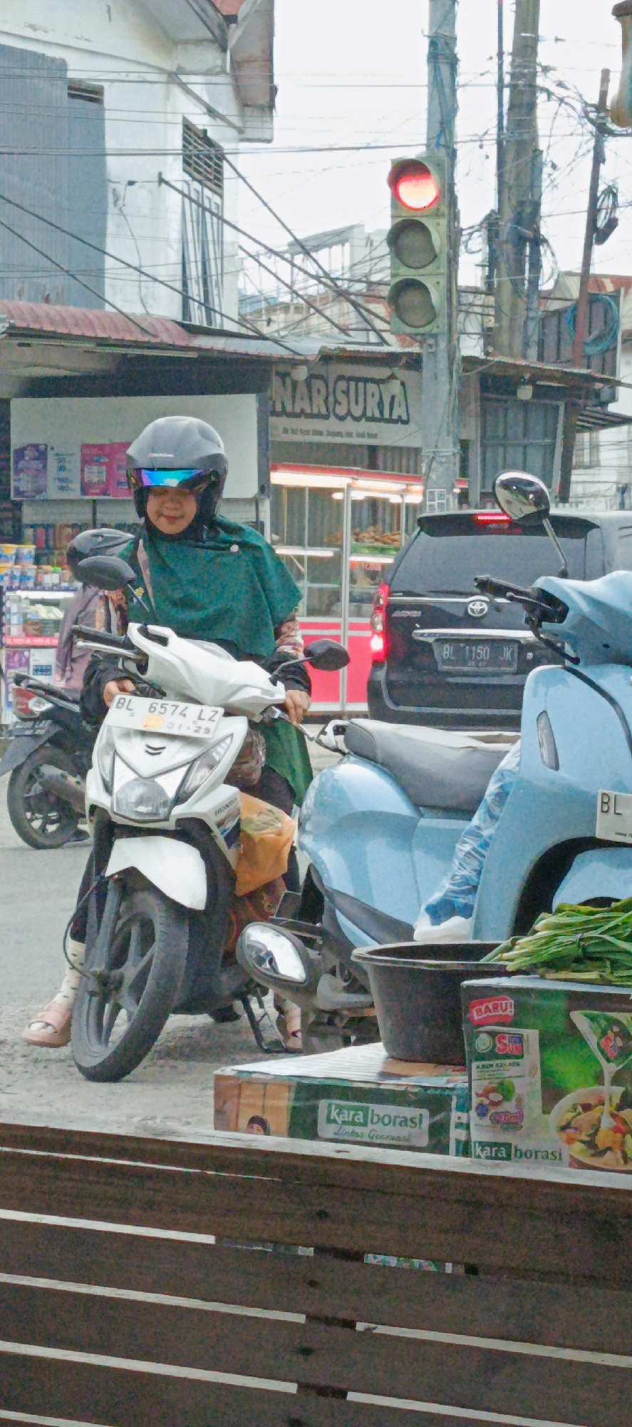 In the city of Garut, Indonesia, a woman wearing a green headscarf and a black helmet with a blue visor is stopped on a white motorcycle at a traffic light. The traffic light is red. The woman is looking forward, and her hands are on the handlebars. The motorcycle has a license plate that reads "BL 6574 LZ". In the background, there is a black car with a license plate that reads "BL 1150 JK". There are also several shops and buildings visible. In the foreground, there are some boxes with produce.