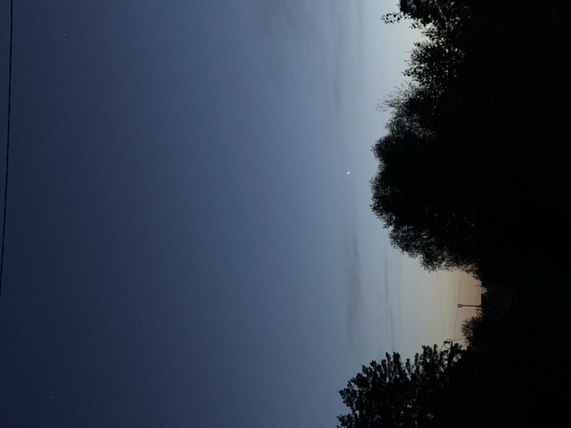 The image shows a night sky, likely during twilight. The sky transitions from a dark blue on the left to a lighter gradient towards the right, suggesting the presence of the sun or moon just below the horizon. There are a few faint stars visible in the darker areas of the sky. The top and bottom of the frame are obscured by silhouetted trees, their dark shapes framing the sky. On the right, utility poles and wires can be seen silhouetted against the lighter portion of the sky. The location is potentially in Chaikovsky, Russia, based on the prompt. Specific details about people, subjects, interactions are not visible in the image.