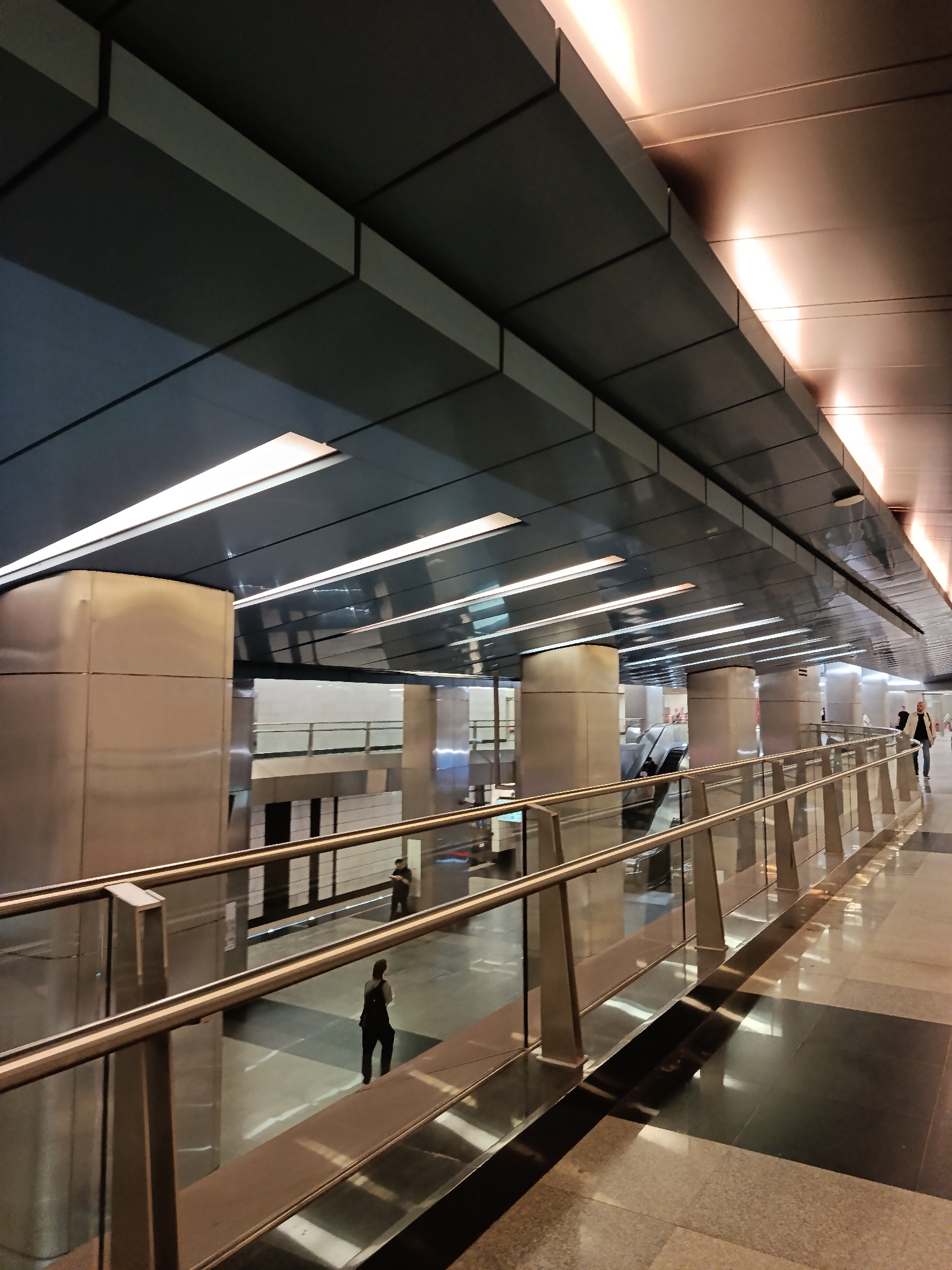 The image shows the interior of a modern metro station in Moscow, Russia. The architecture features a high ceiling constructed with dark gray rectangular panels interspersed with long, linear fluorescent lights. The station has polished floors that reflect the ambient light.
A stainless steel railing runs along an elevated walkway, providing a view down to a lower level. Large, cylindrical pillars, also stainless steel, support the structure. An escalator can be seen in the background.
Several people are present: one individual stands on the lower level, another walks away in the same area, and a third person is walking near the end of the walkway on the right.